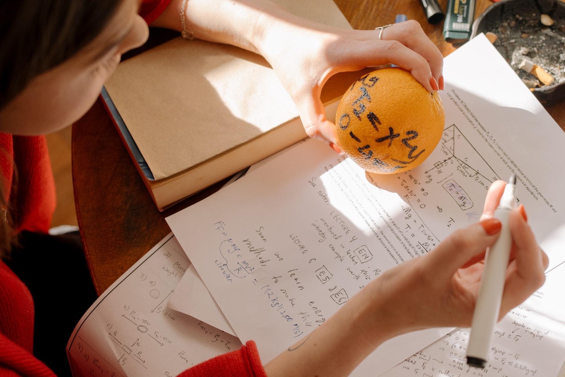 Caderno de matemática sobre a mesa. Sobre ele, duas mãos. Enquanto uma segura um lápis, a outra segura uma bolinha laranja com números estampados em preto.