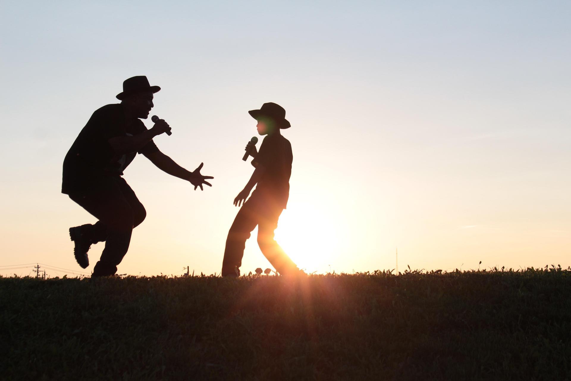 Campo de grama ao entardecer. Na foto aparece basicamente só a silhueta dos integrantes que são um homem e um menino cantando com o microfone nas mãos, um de frente para o outro.
