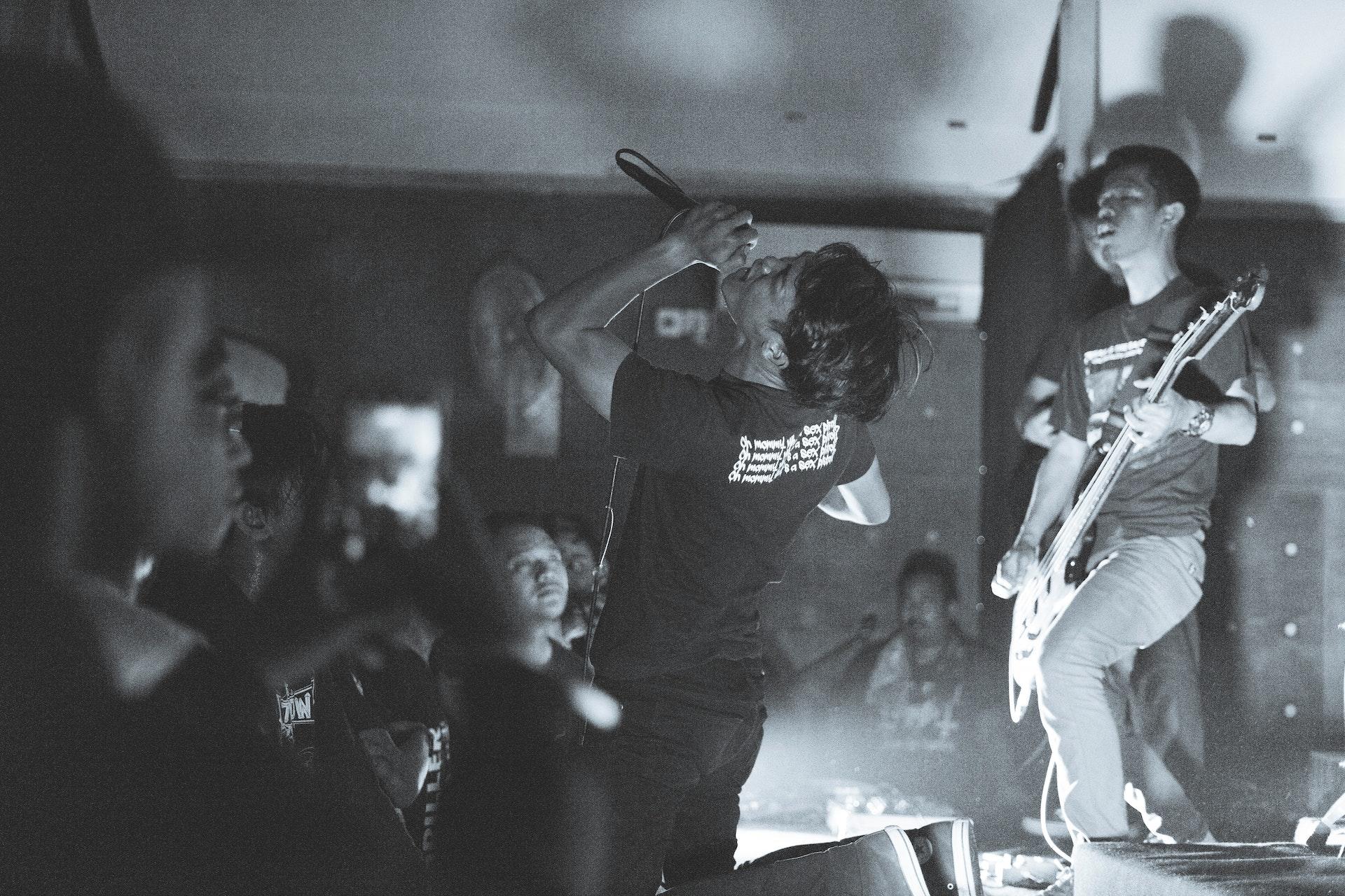 Foto preta e branca. Banda se apresentando em um bar. Um integrante tocando guitarra e o outro cantando com a cabeça para cima. O público está assistindo e filmando com celulares.