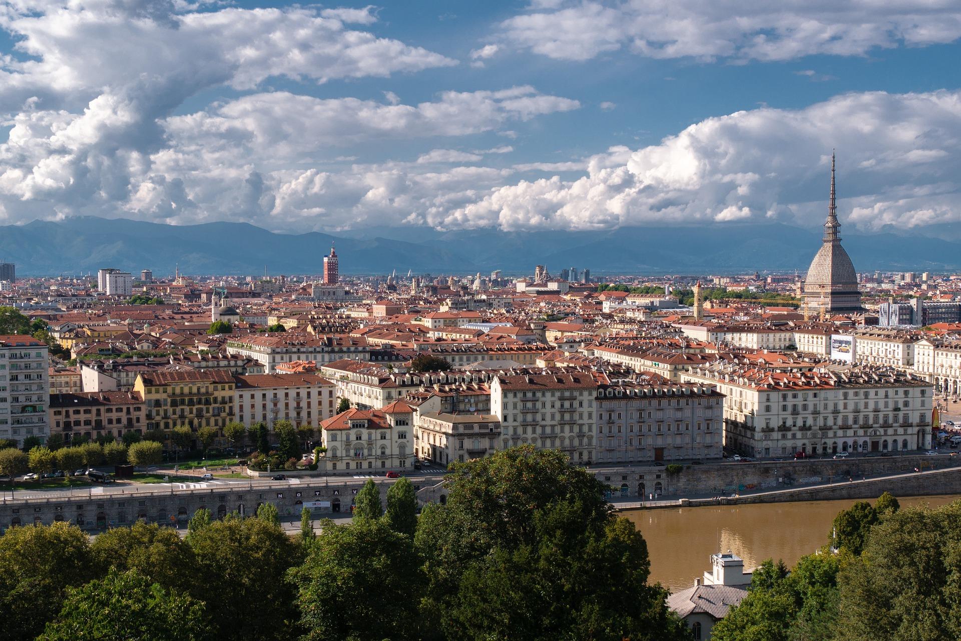Vista panorâmica da cidade de Turim, onde o céu está nublado, mas percebe-se alguns raios de sol. A foto mostra diversas construções e árvores à frente.