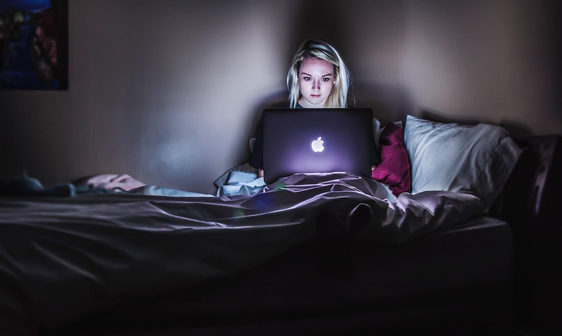Moça loira em um quarto escuro sentada na cama com um computador Apple no colo. É possível ver a claridade da tela refletindo em seu rosto.