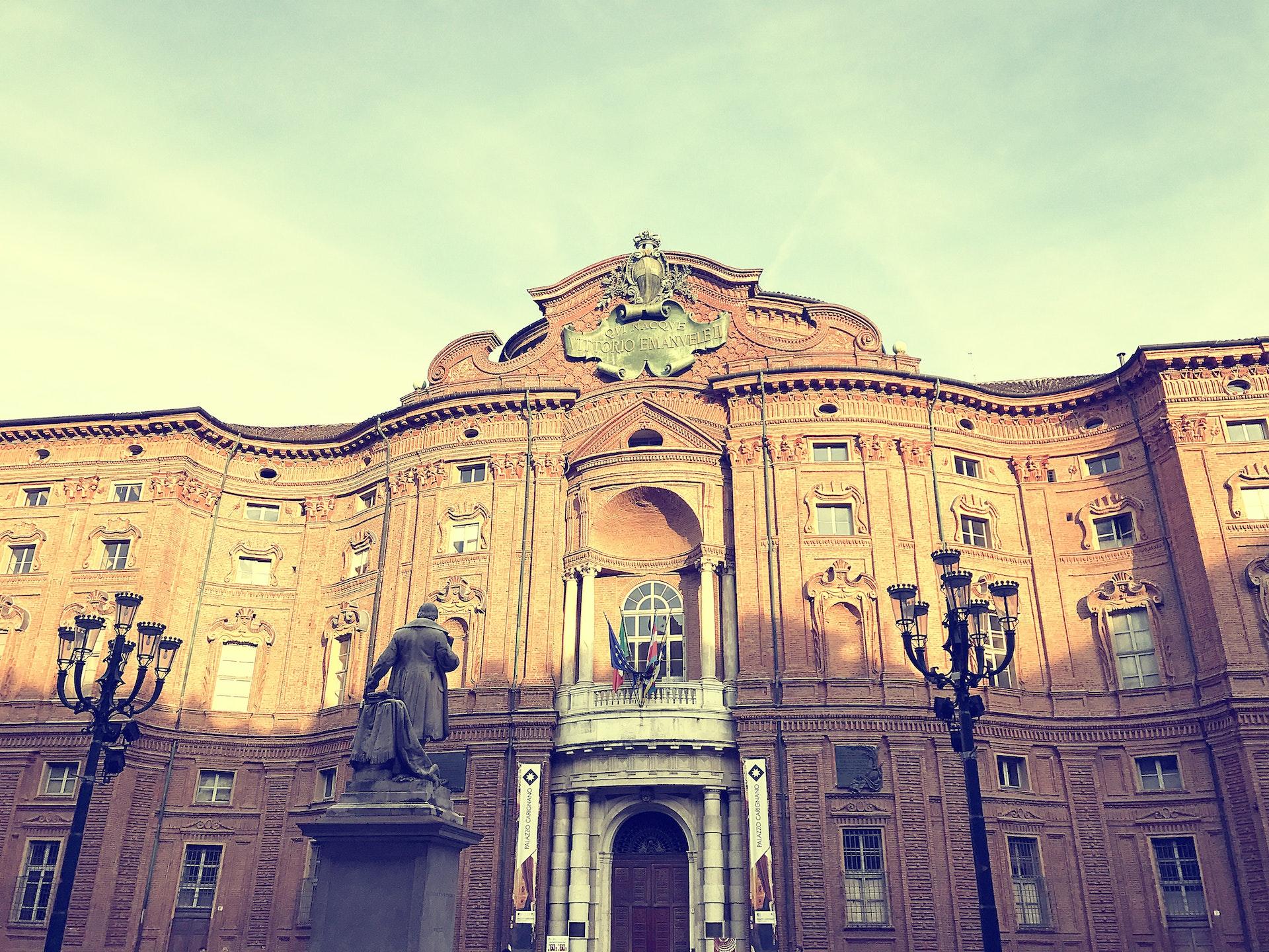 Edifício histórico em Turim na Itália. Em bege, visto de perto, com raios de sol refletindo. Á frente, é possível ver luminárias nos modelos antigos e um monumento.