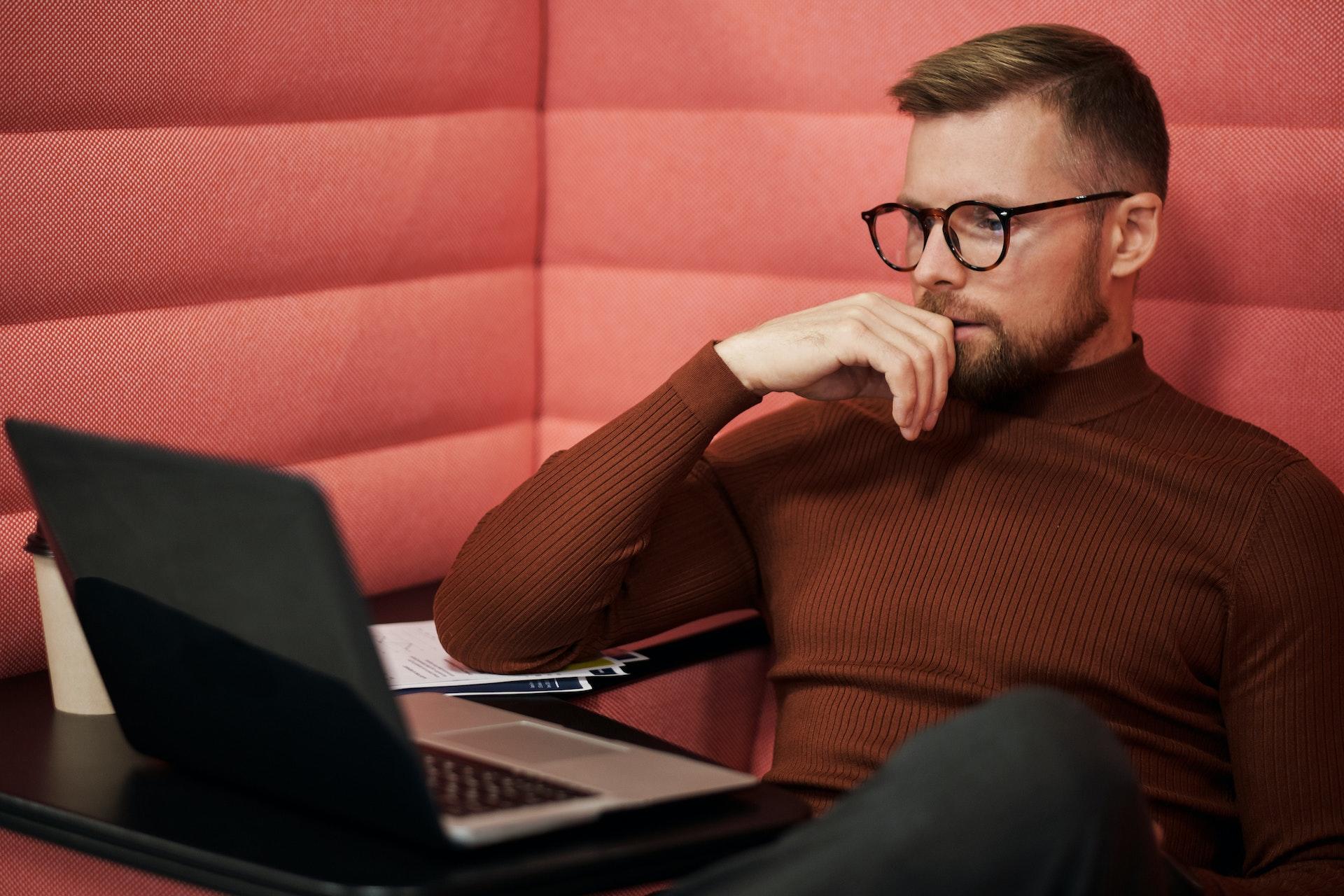 Homem sentado em um sofá cor de rosa, com um computador à sua frente. Ele usa óculos e um suéter marrom. A mão apoiada na boca sugere que está atento ao que lê. Fonte: Pixabay