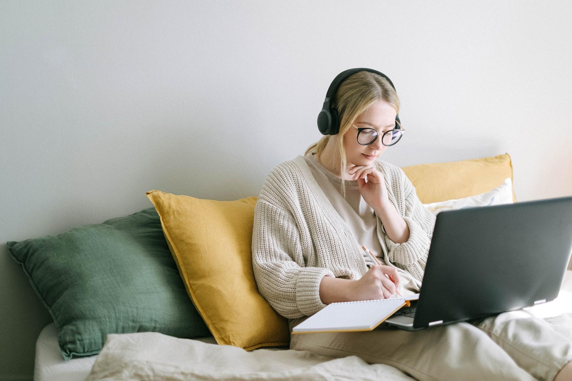 Mulher loura, usando óculos e headphones, Ela está sentada em um sofá amarelo, com um computador no colo e um caderno, no qual está fazendo anotações.