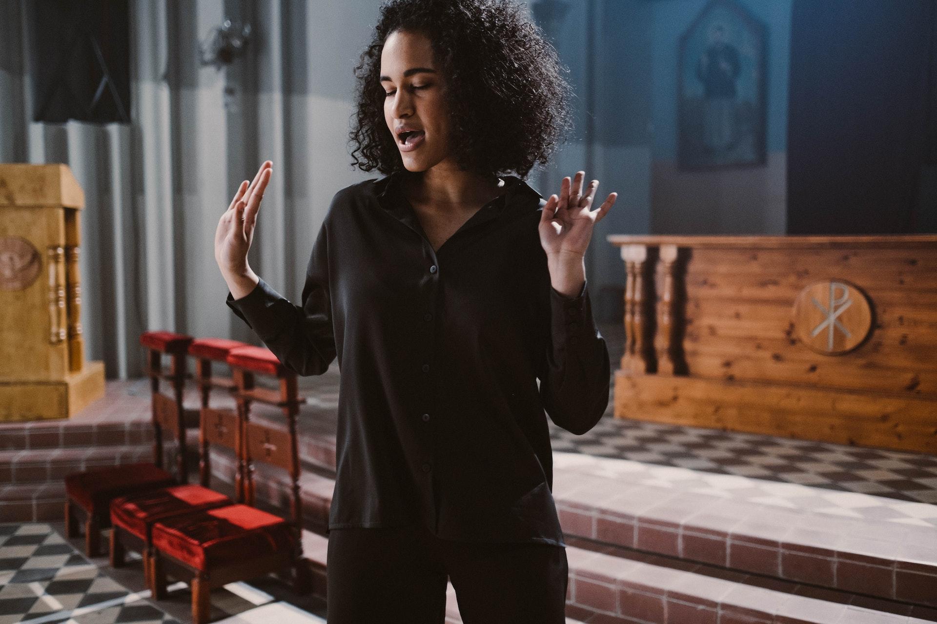 Mulher morena dos cabelos cacheados na altura dos ombros, com camisa e calça preta, dentro de uma igreja, cantando de olhos fechados e mãos semi-elevadas.