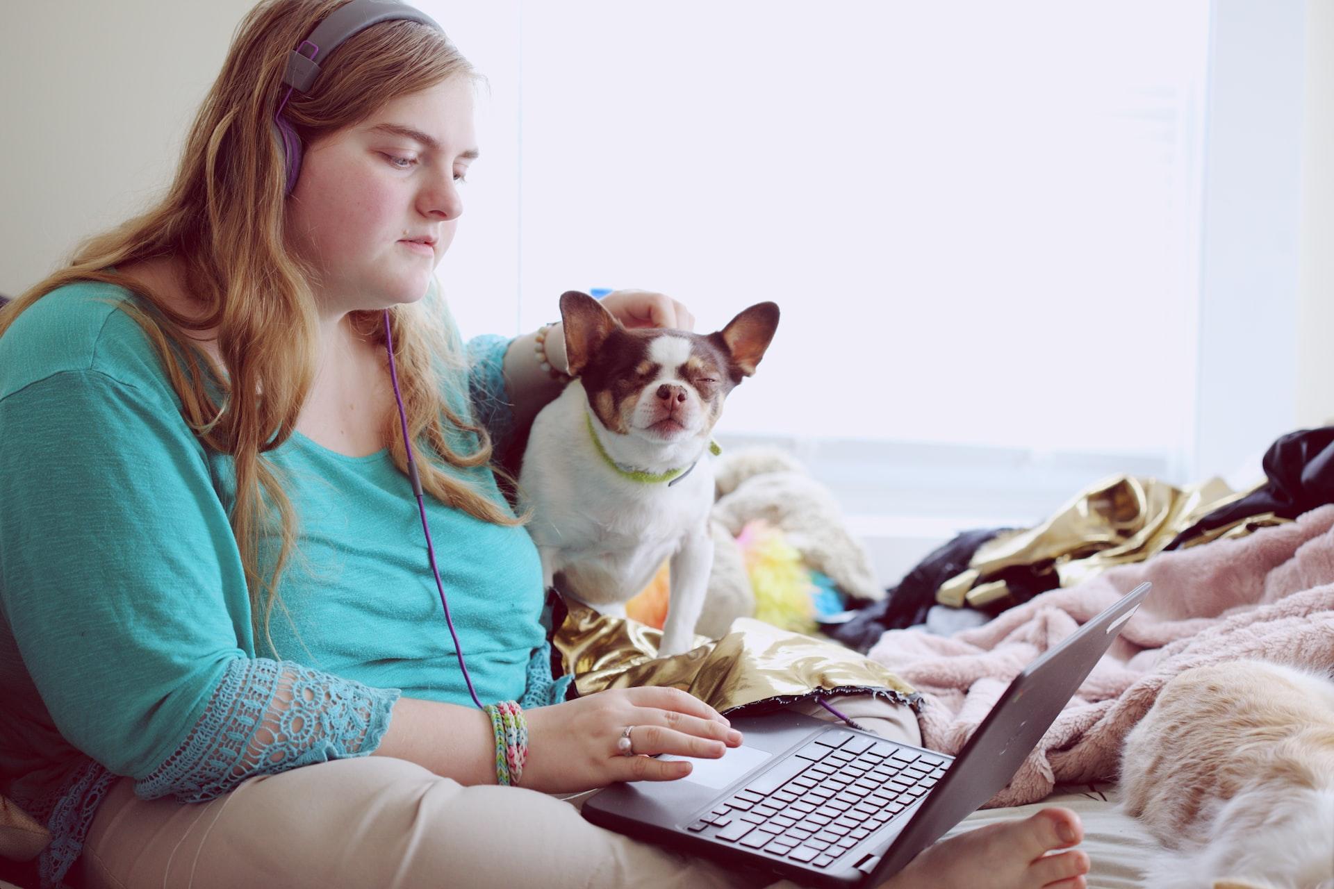 mulher sentada na cama vendo algo em seu computador e fazendo carinho em um cachorro