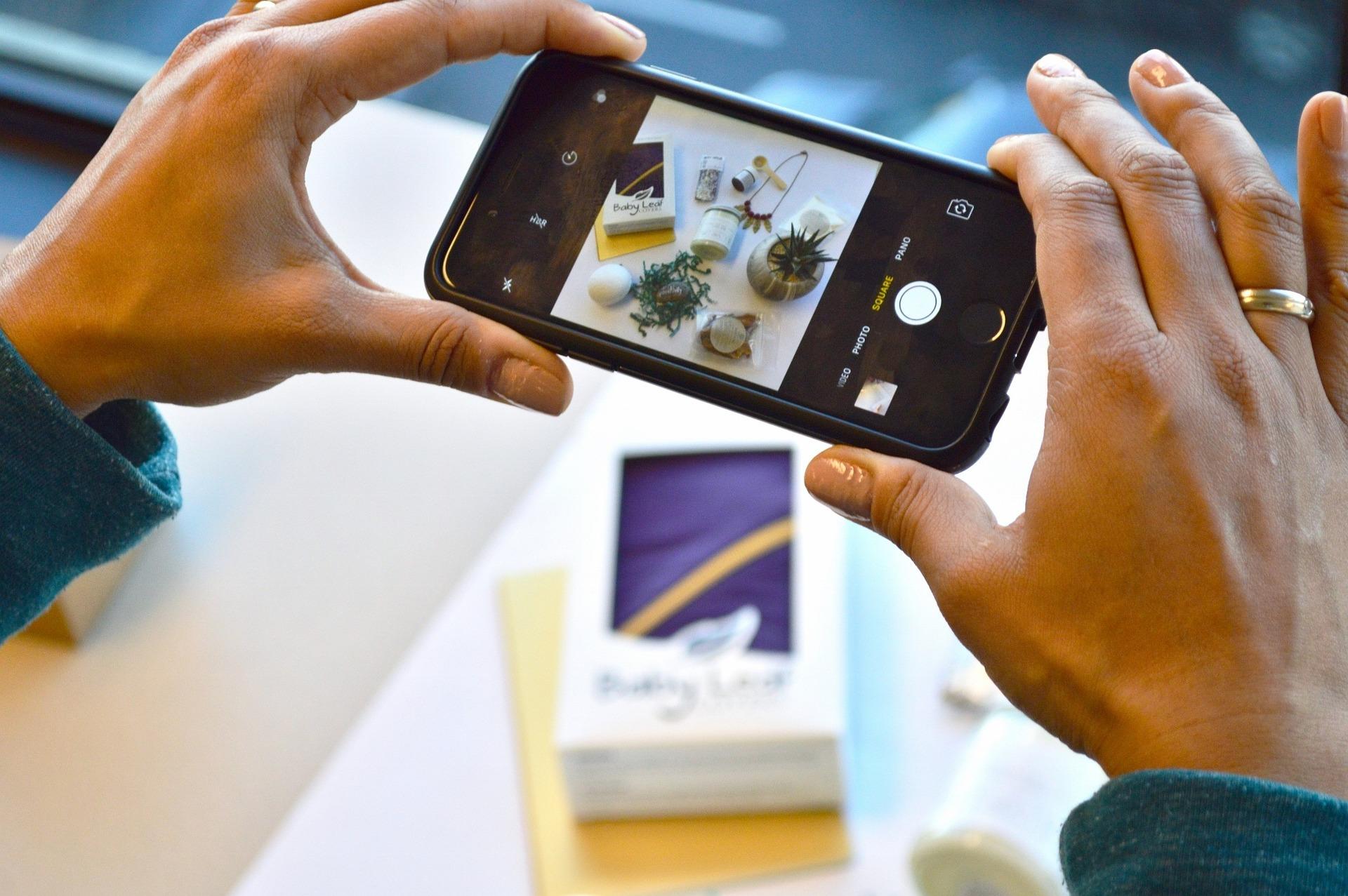 Mãos femininas segurando um celular para tirar fotos de alguns produtos em uma mesa com um fundo branco.