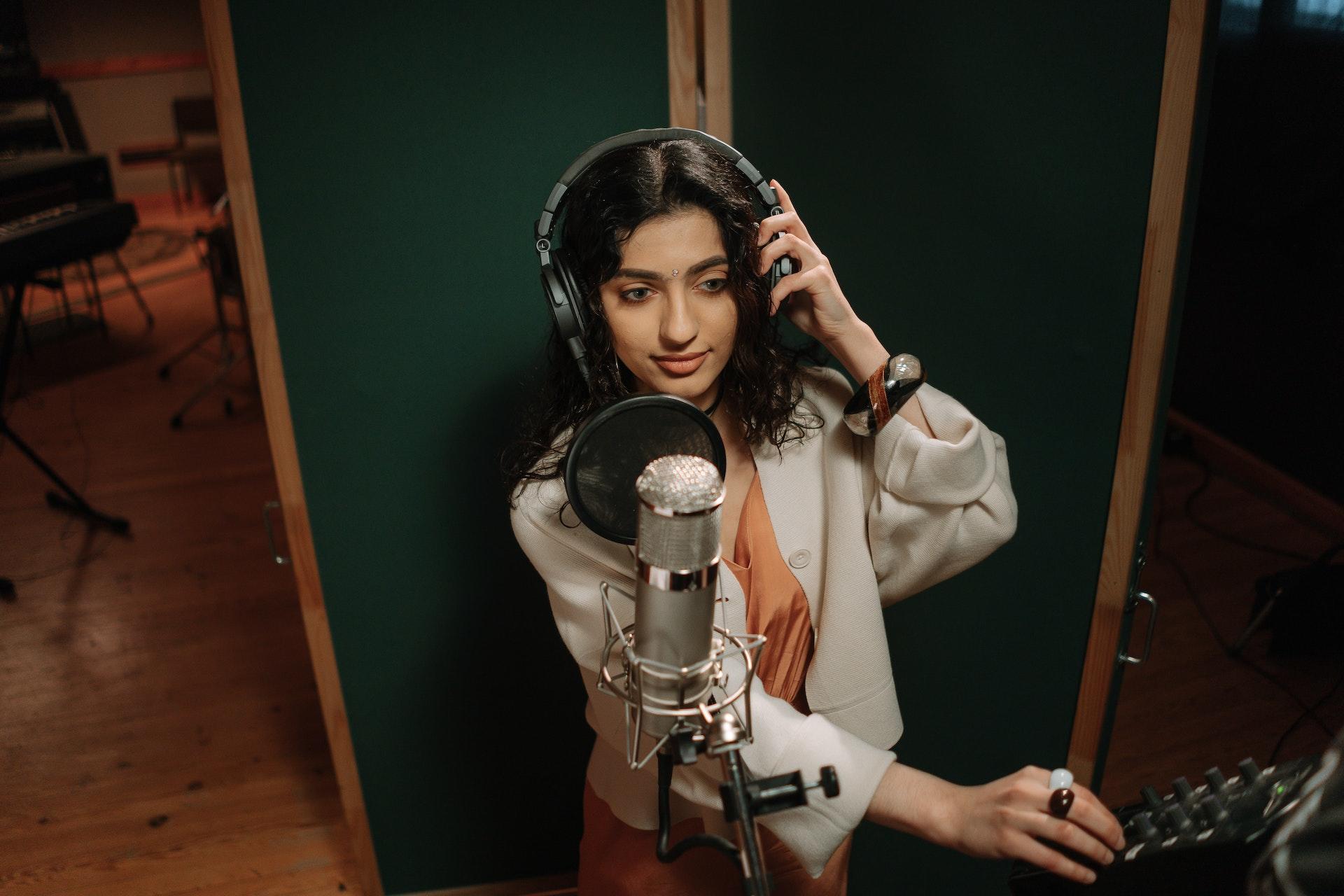 Moça branca com cabelos castanhos ondulados. Usando um headset e mexendo em um painel de rádio, com um microfone prata próximo. Ao fundo, é possível ver pés de instrumentos.