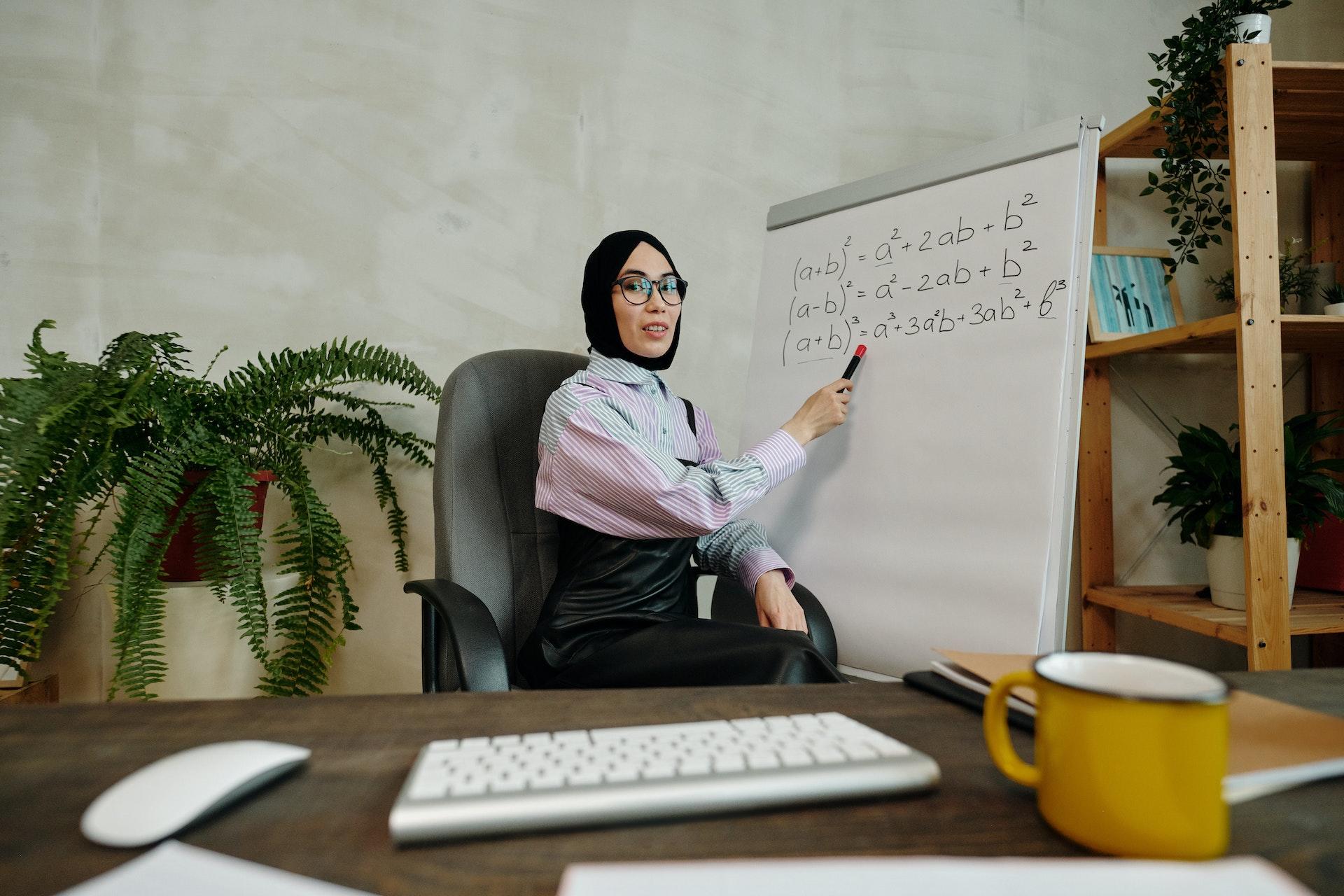 Mulher com hijab preto, sentada em uma cadeira, apontando para uma lousa, na qual há algumas fórmulas matemáticas. Á frente dela, podemos ver um teclado sobre uma mesa, indicando que a imagem da foto sugere o que o aluno vê pela tela do computador.