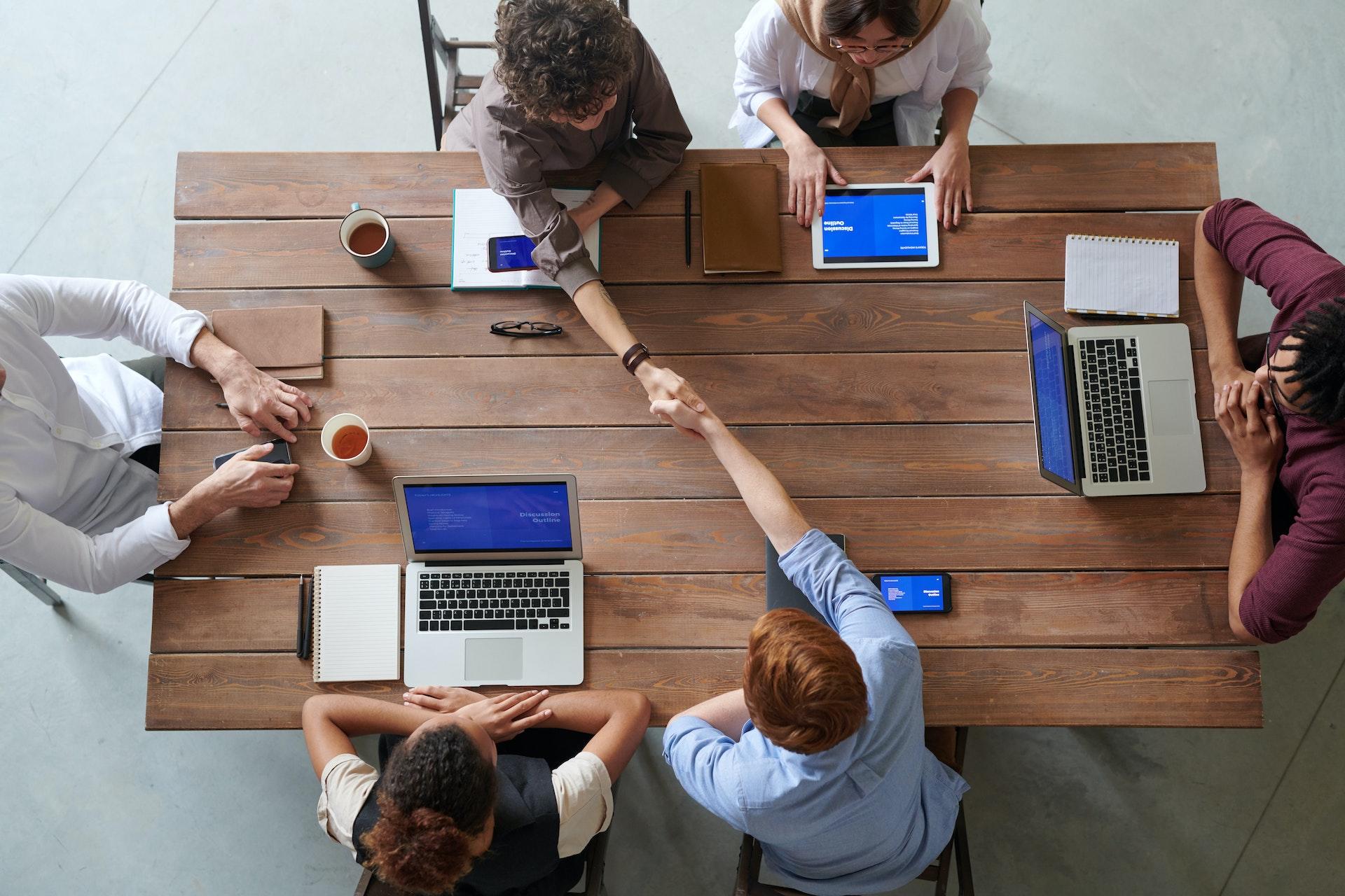 Bancada vista de cima com 6 computadores. Em frente a cada um, há uma pessoa sentada. Mãos dadas e materiais