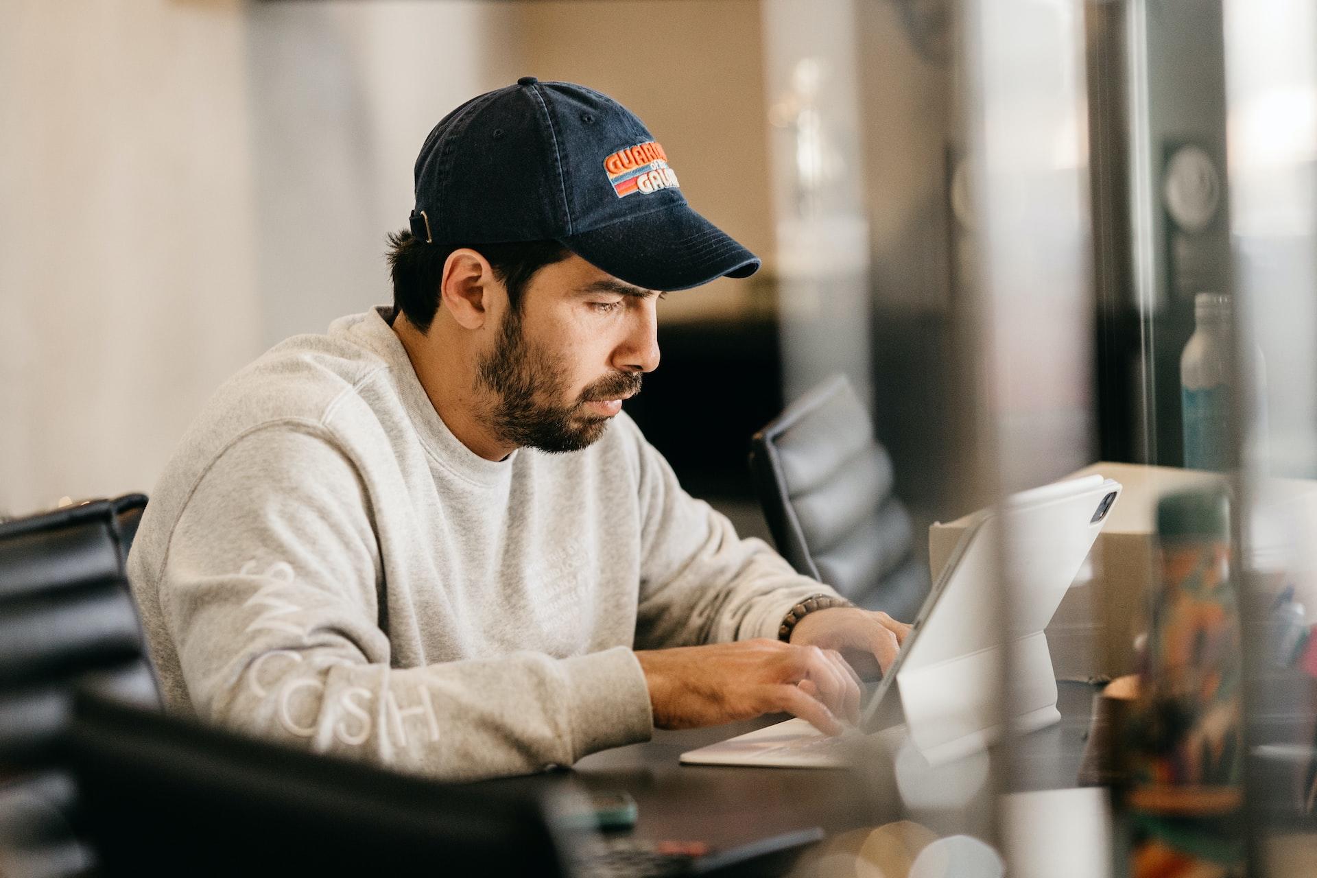 Homem de boné, sentado em frente a um computador com mãos no teclado e olhar concentrado. É possível ver que ele está dentro de um escritório. Fonte: Unsplash