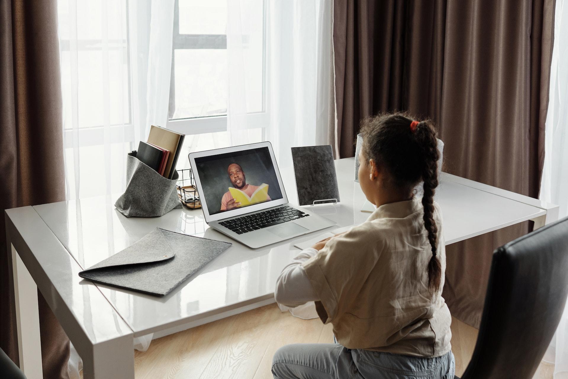 Menina sentada em uma escrivaninha em seu quarto, estudando online. Ela tem uma trança no cabelo e está de costas para a câmera. Sobre a mesa, há o computador e mais alguns materiais escolares.