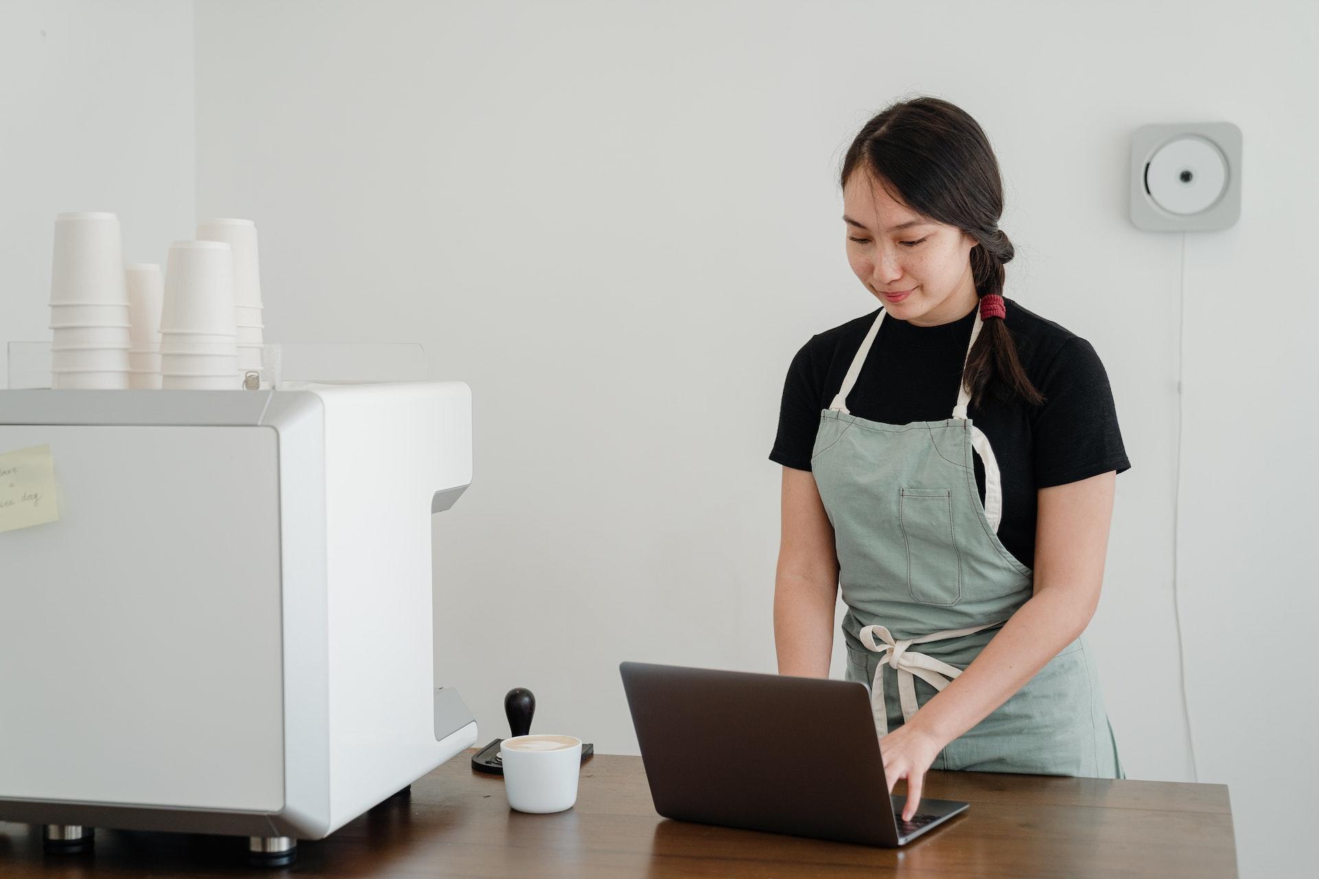 Computador sobre uma mesa de costas para a câmera. De frente, há uma moça em pé atrás da mesa, mexendo no computador. Também há um eletrodoméstico branco com copos descartáveis sobre ele.