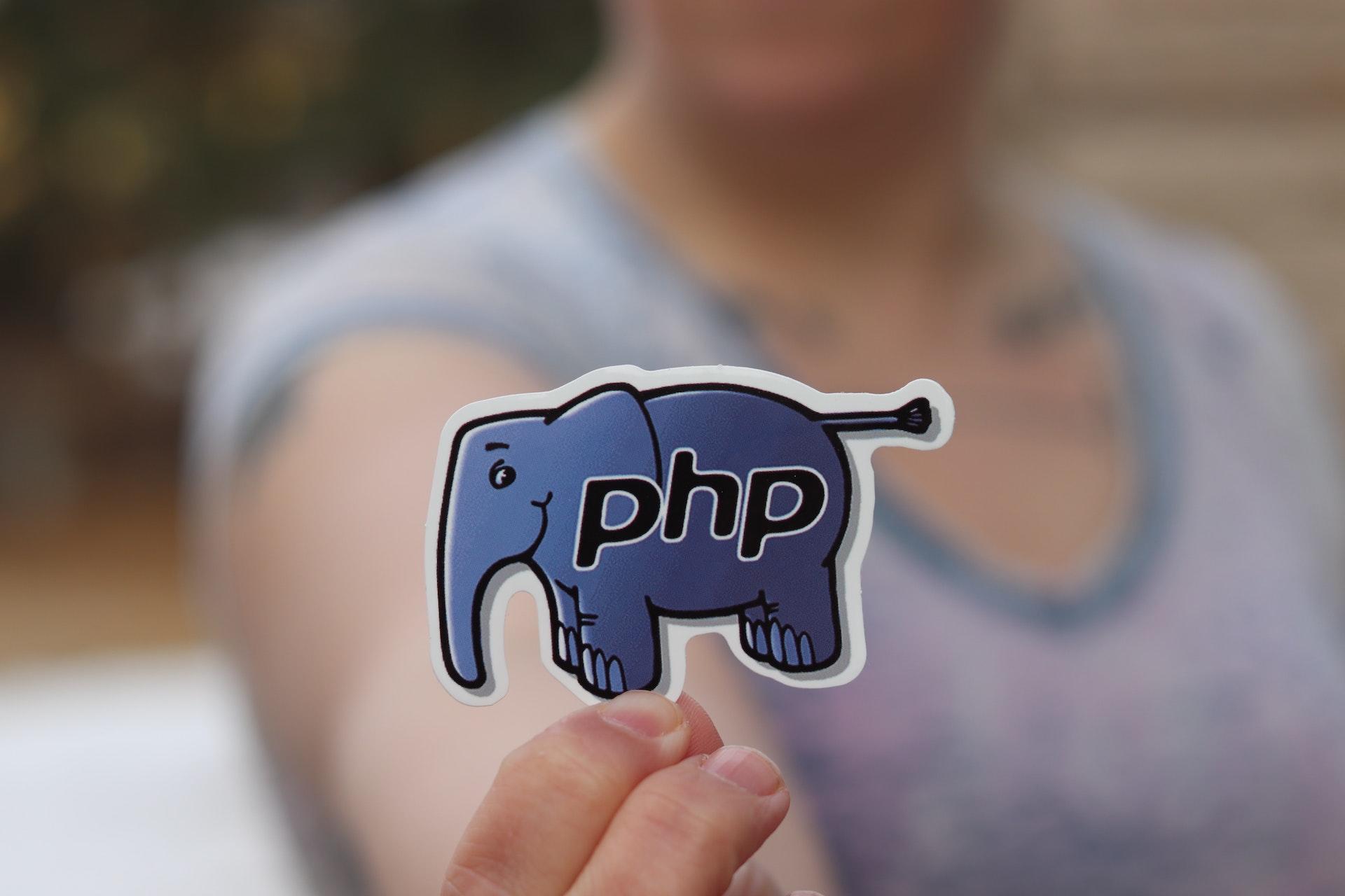 Etiqueta em formato de elefante com a sigla "PHP". É a única parte nítida da foto, bem no destaque. Na imagem desfocada, é possível ver o pescoço, braço e mão da pessoa que segura a etiqueta.
