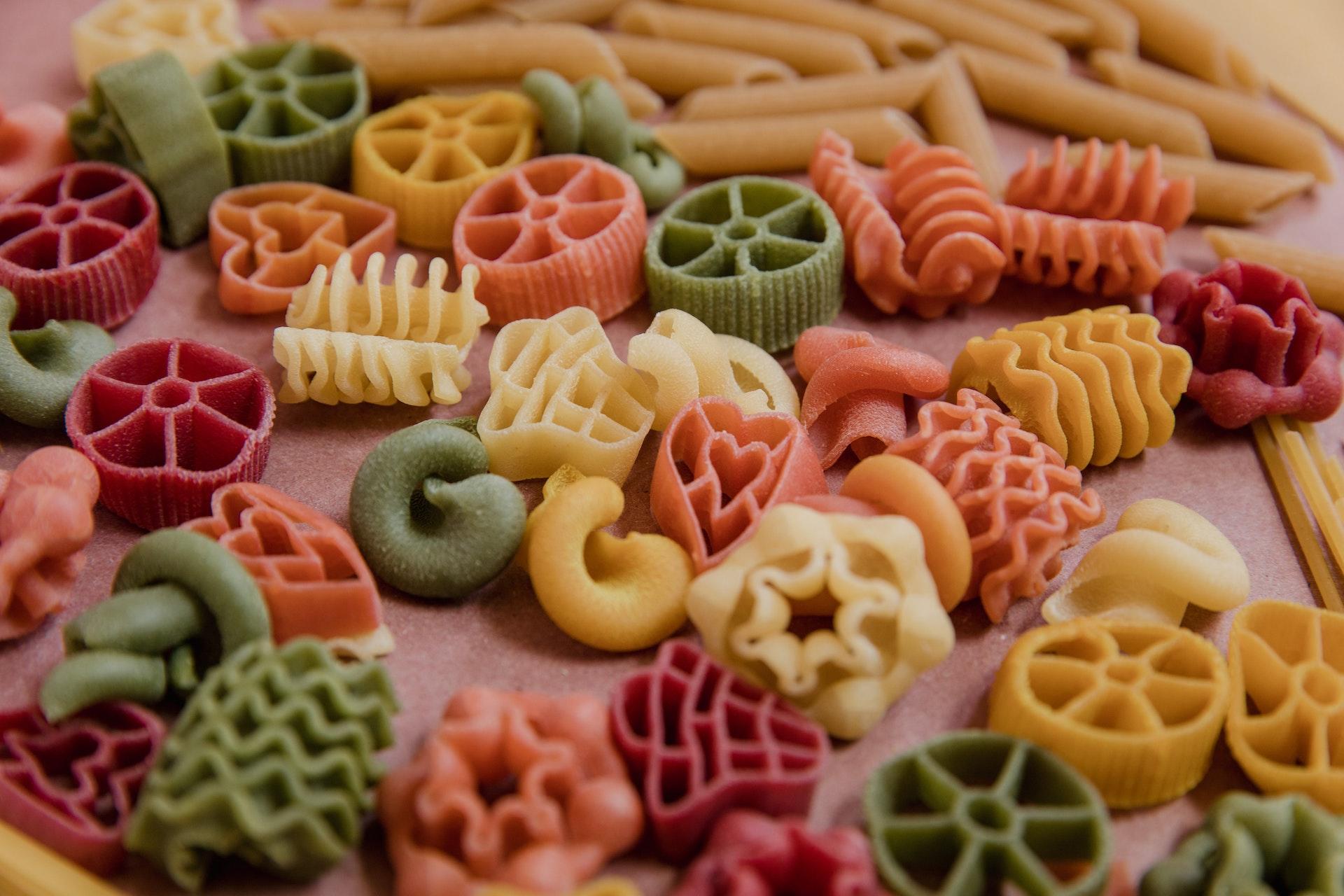 Macarrão em várias cores e formatos, crus, colocados sobre um fundo avermelhado.