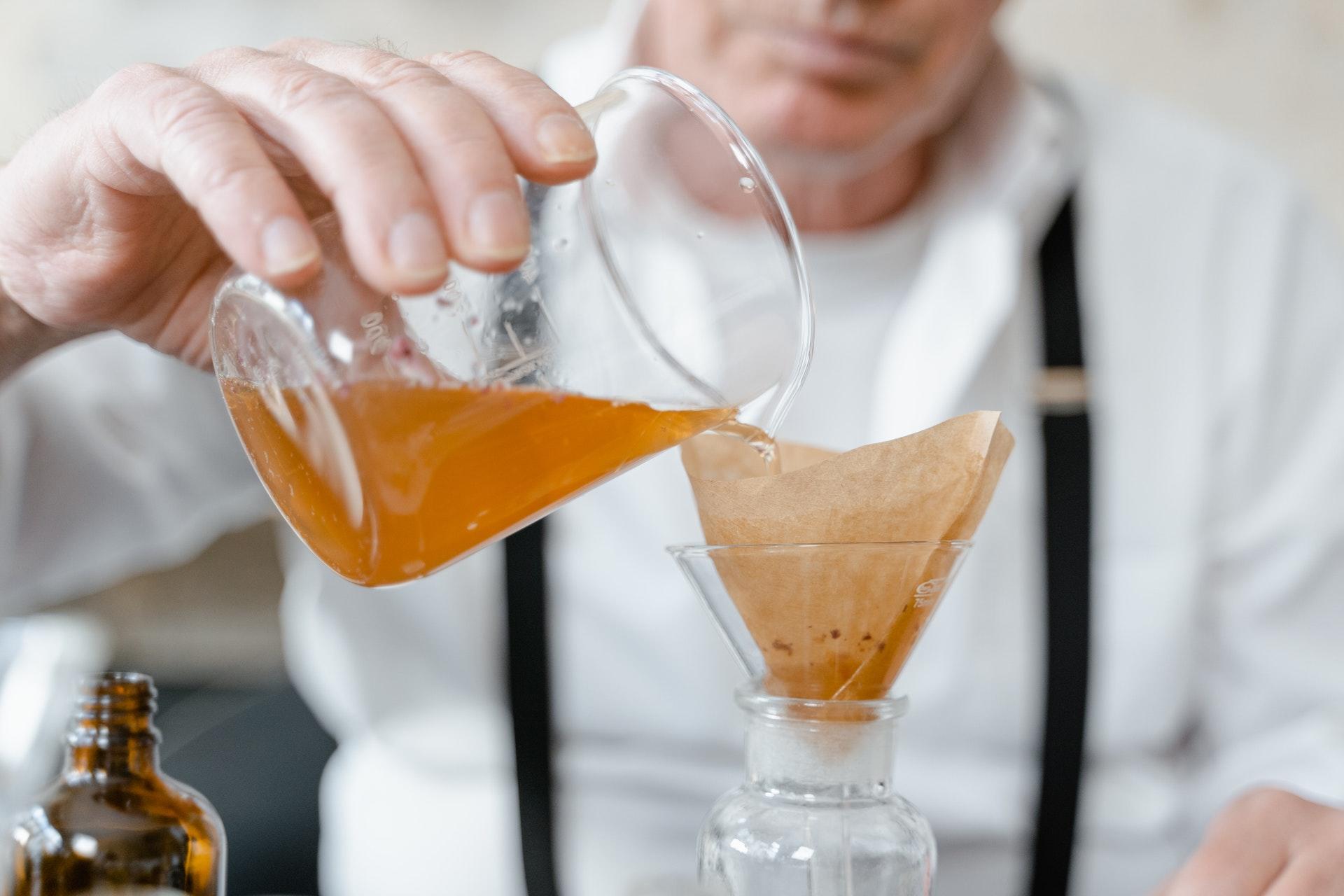 O foco da foto são as mãos de um homem branco, de roupas brancas separando uma mistura de óleo com um coador de papel e uma garrafa em laboratório. Não é possível ver o seu rosto. 