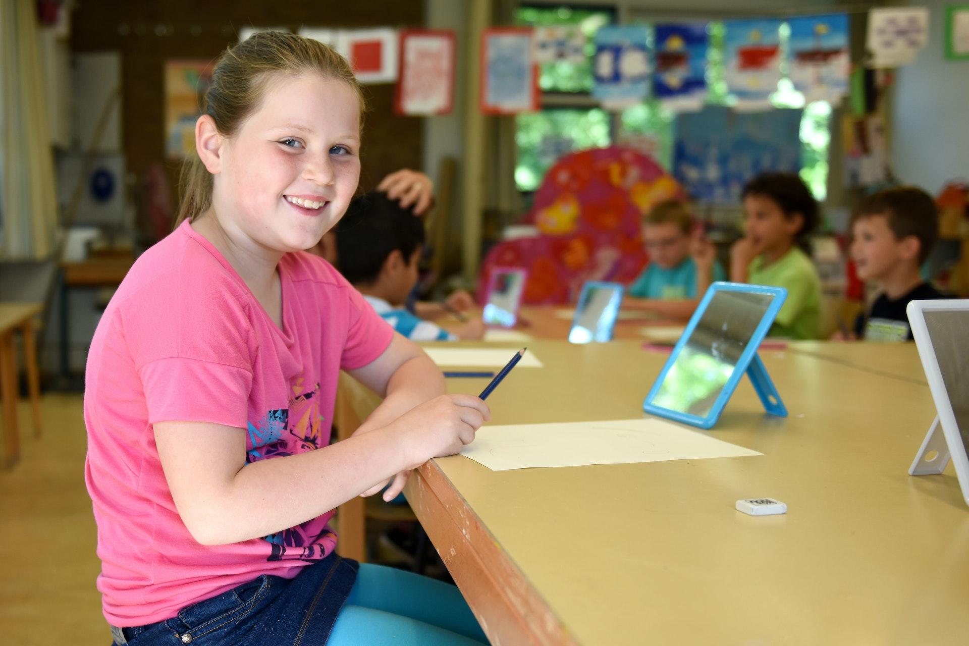 Menina sentada em um laboratório de escola, decorado com cores alegres, de frente com um tablet. Ela usa rosa e tem um sorriso no rosto.