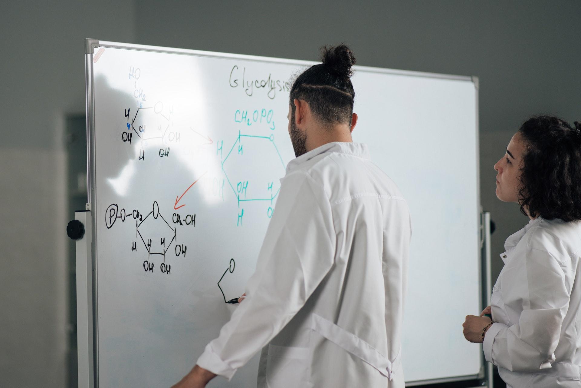 Professor com jaleco branco desenhando fórmulas e estruturas de química em uma lousa branca e sendo observado por uma mulher também de jaleco branco.