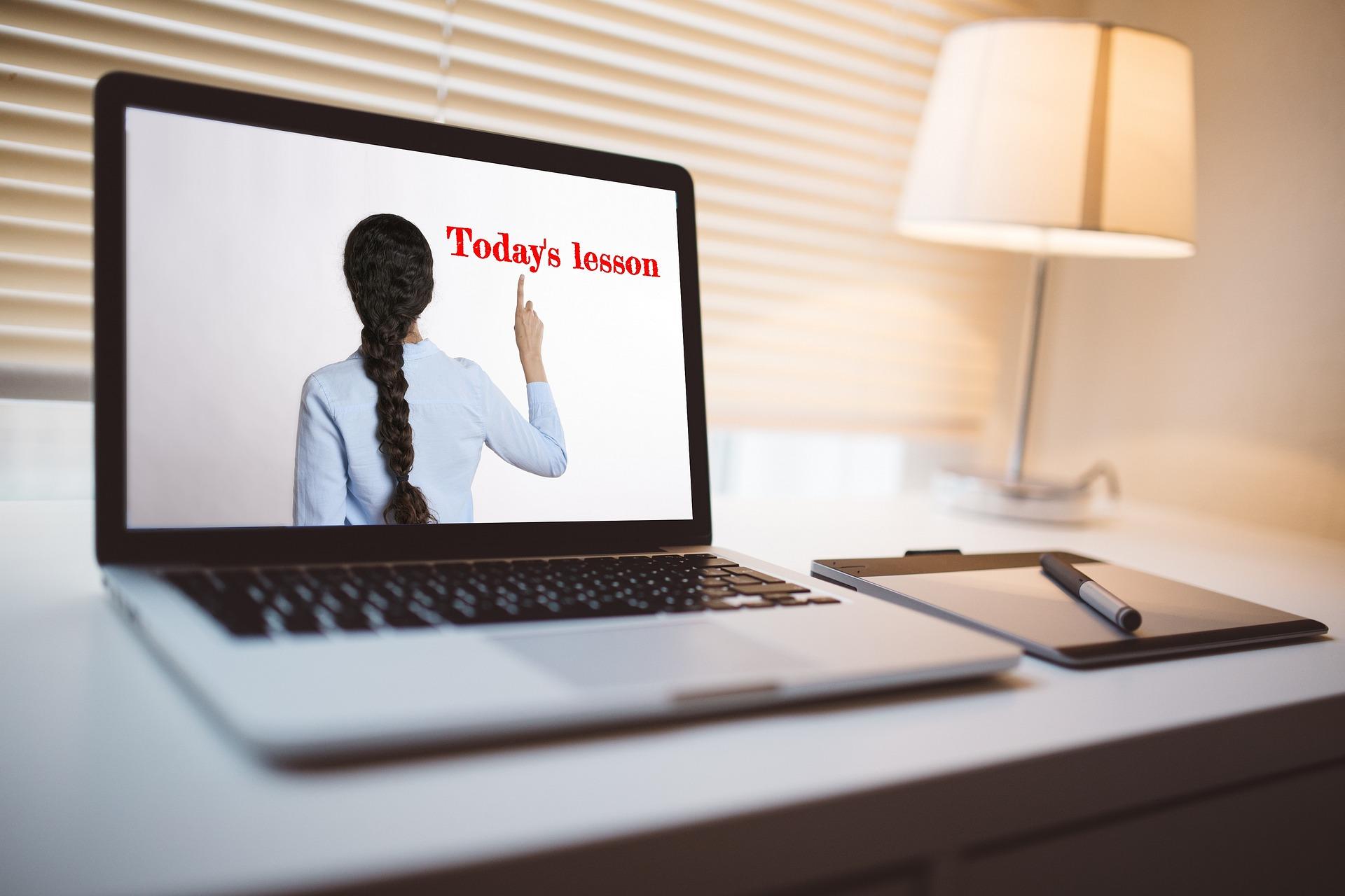 Laptop sobre a mesa. Ao lado, um bloco de anotações e uma caneta. Na tela, há uma professora de costas, com blusa branca e trança no cabelo. Ela aponta para a frase: "Today's lesson".
