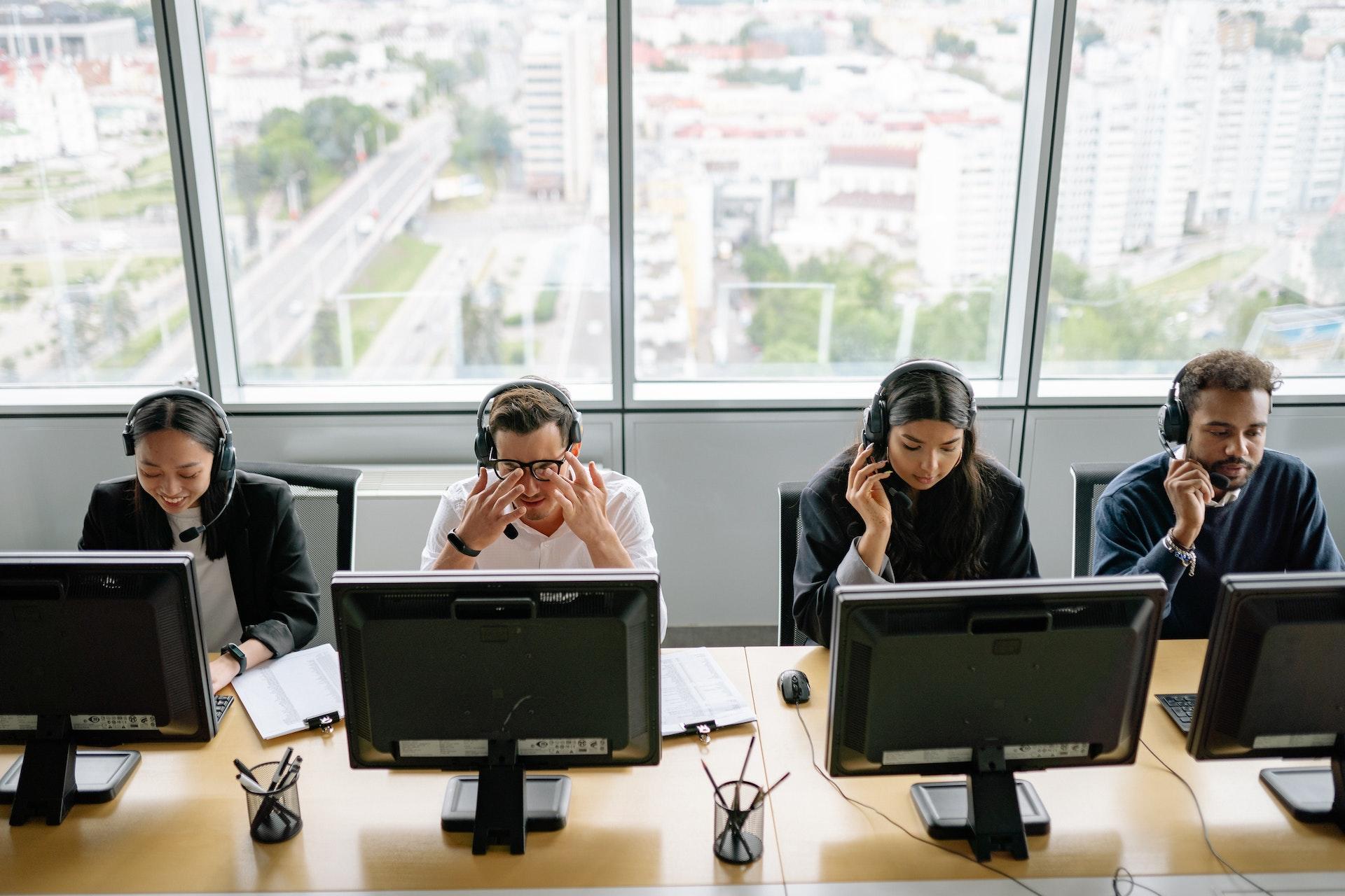 Pessoas sentadas diante de computadores. Na foto, aparecem 4 pessoas sentadas de frente para a câmera (os computadores estão de costas). 