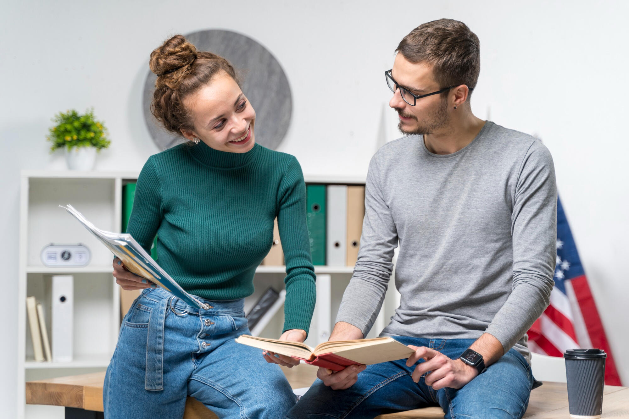 Um homem e uma mulher, segurando livros de inglês, e sentados em cima da mesa, praticando pronúncia em inglês.