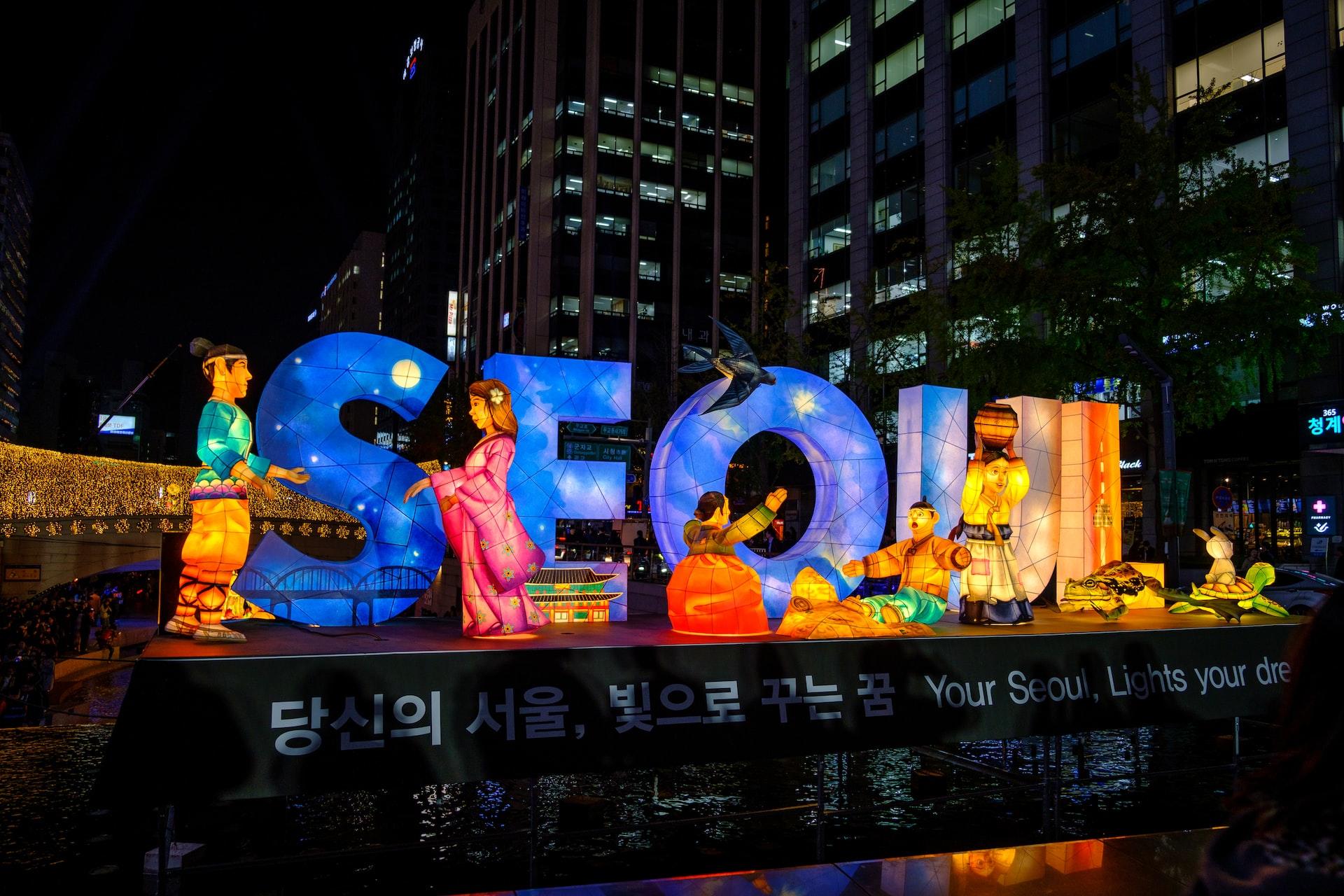 Letras iluminadas na palavra Seul em inglês, com destaque de personagens à noite.