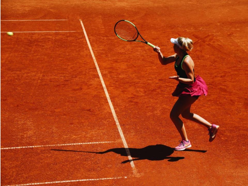Mulher com roupa esportiva de tênis, de pé em uma quadra de tênis, segura a raquete com a mão direita após rebater a bola amarela que é vista no canto esquerdo da imagem.