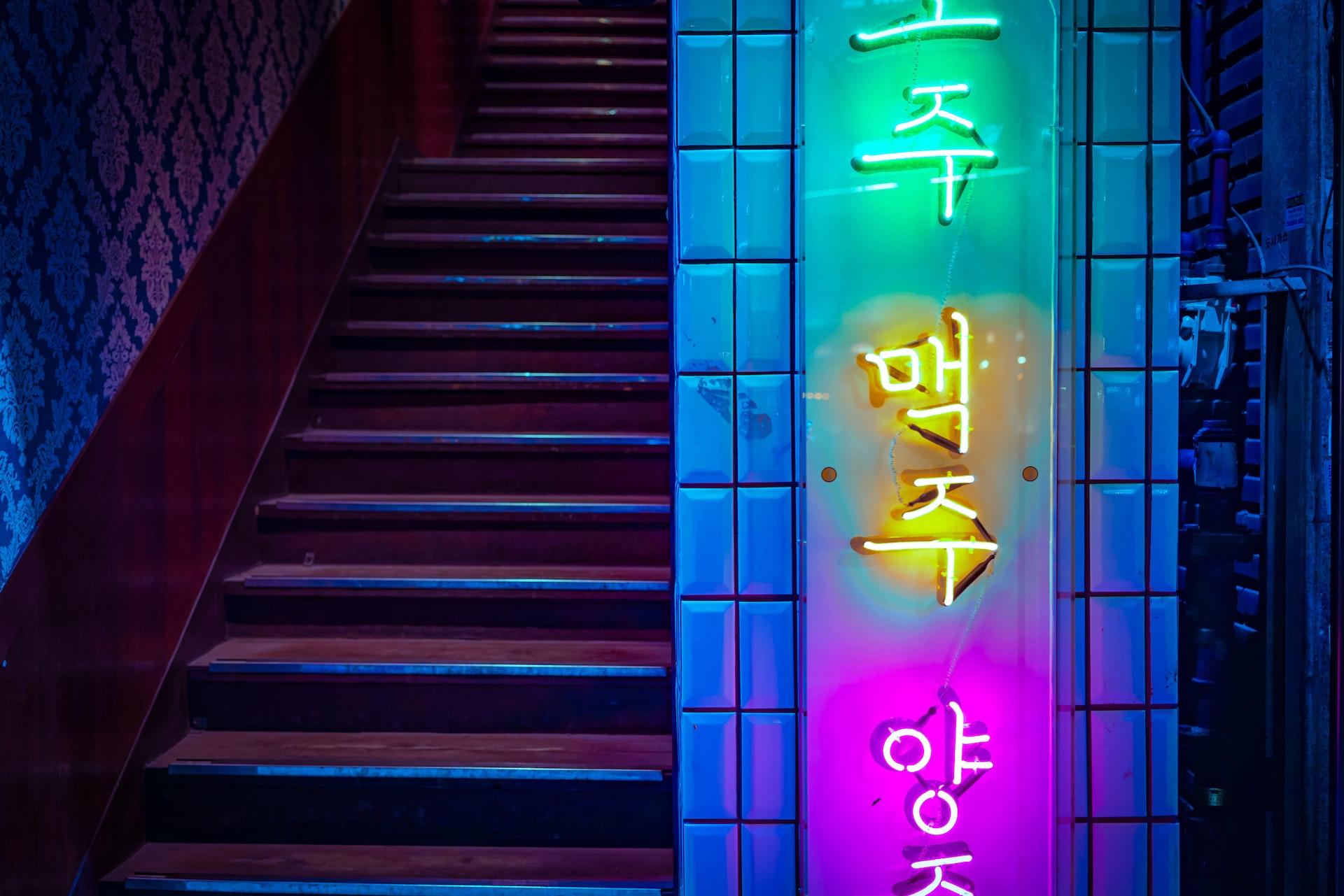 Letreiro luminoso com letras coreanas, ao lado de um escadaria com reflexo de luzes.