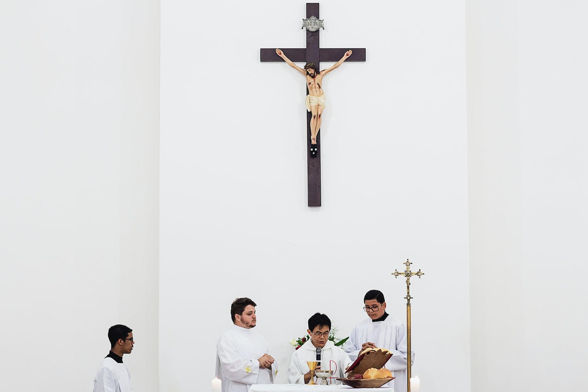 Fragmento do altar de uma missa católica, com padres e seus auxiliares. Ao fundo, acima, pode-se ver uma imagem pendurada de Cristo na cruz.
