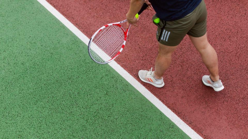 Imagem de um tenista da cintura para baixo segurando a raquete e a bola, mostrando o piso da quadra na cor verde e vermelho separado por uma linha branca.