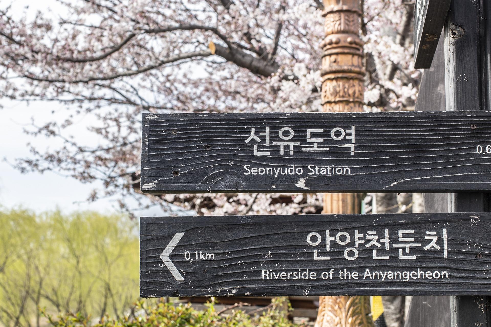 Placas de indicação escritas em coreano. São pretas, escritas em branco e coladas a um poste.