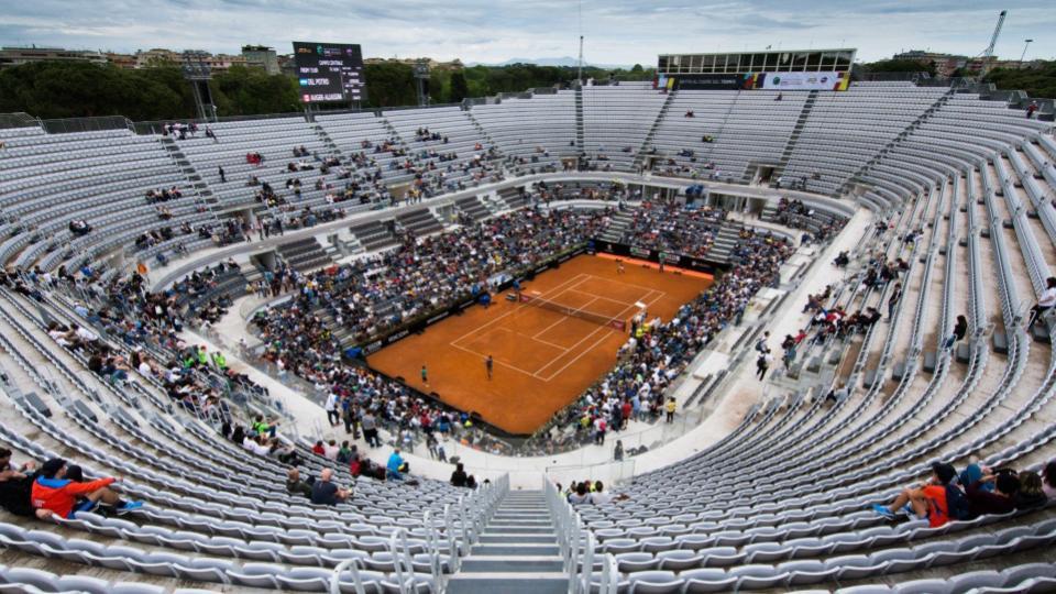 Quadra de tenis sob o sol com pessoas assistindo uma partida