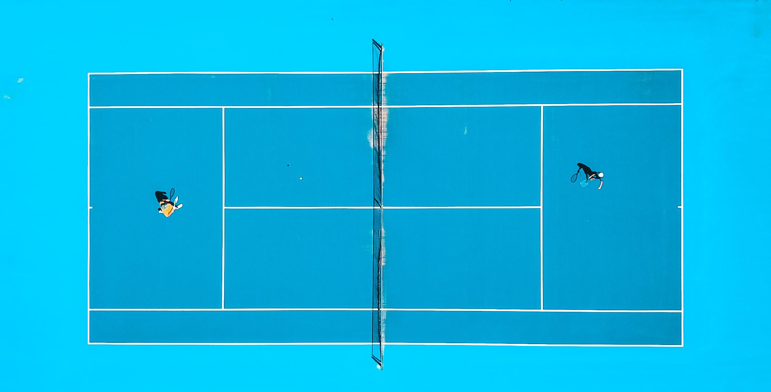 Quadra de tenis com o chão azul de uma visão aérea simples, com dois jogadores um de frente para o outro, sacador e recebedor, separados pela rede. 