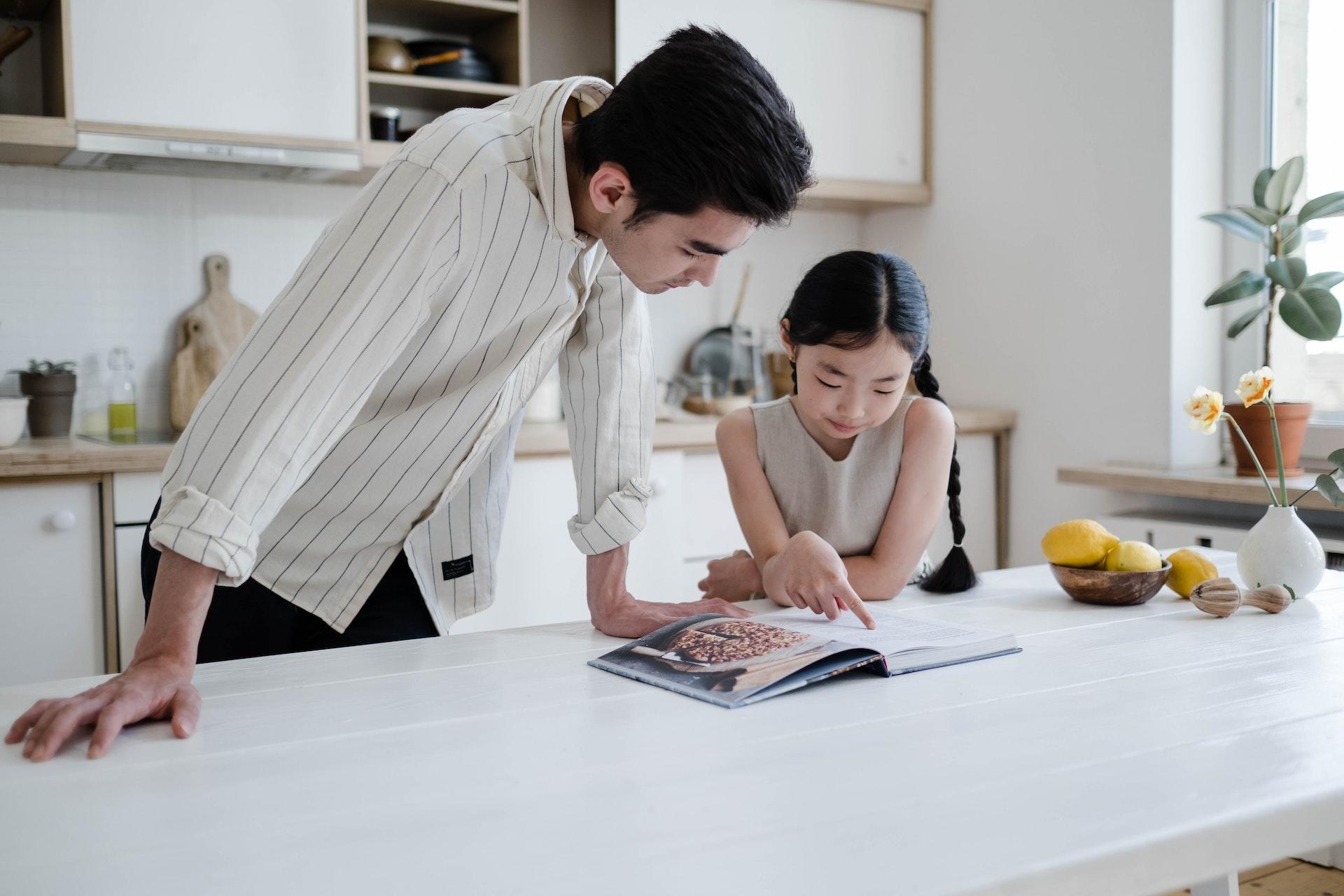 Pai e filha coreanos em uma cozinha bem equipada olhando um livro de receitas.