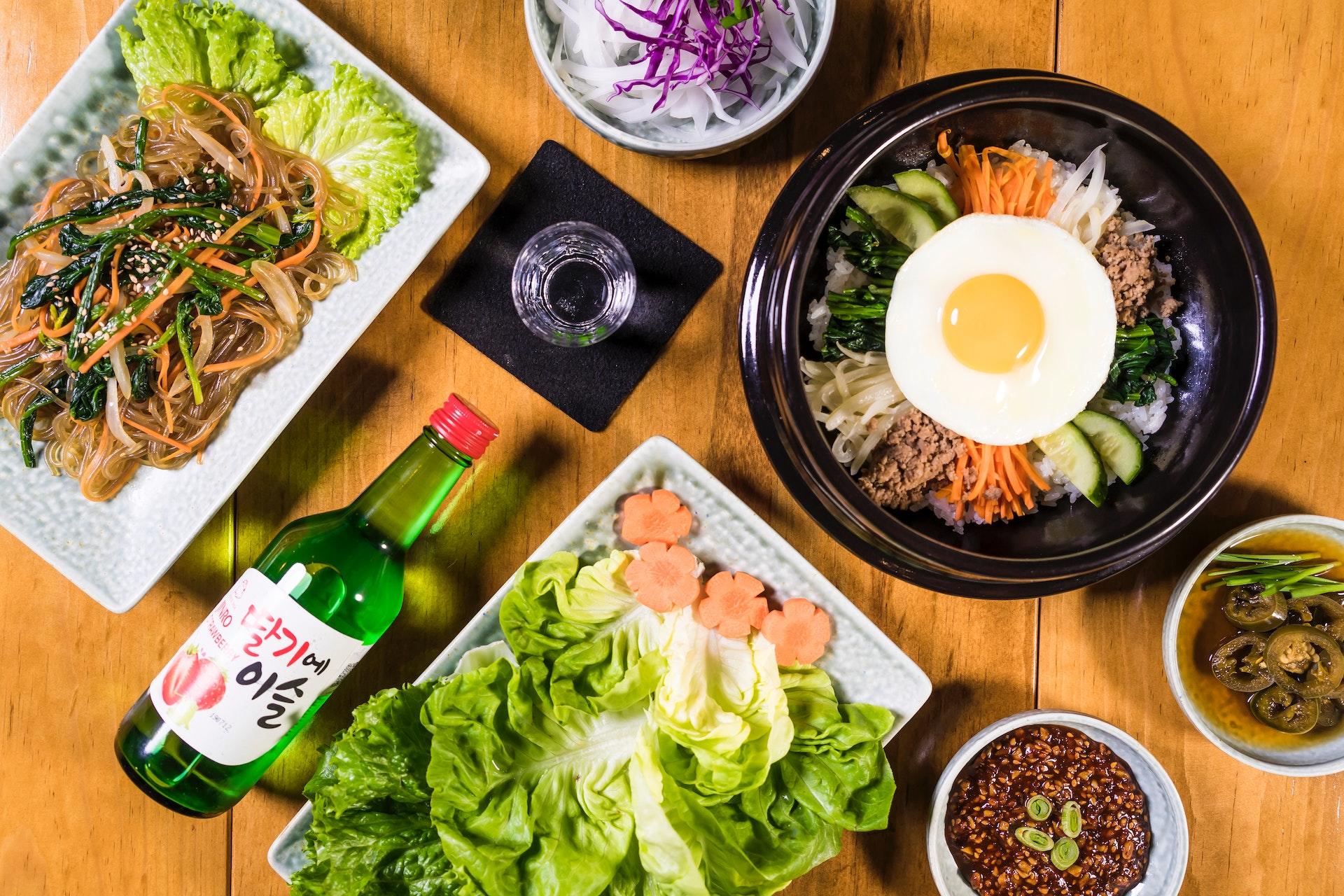 Mesa posta com delícias da culinária coreana. Em destaque, é possível ver o bibimbap com o ovo ao meio. Na mesa também há pratos com vegetais e uma garrafa de azeite.