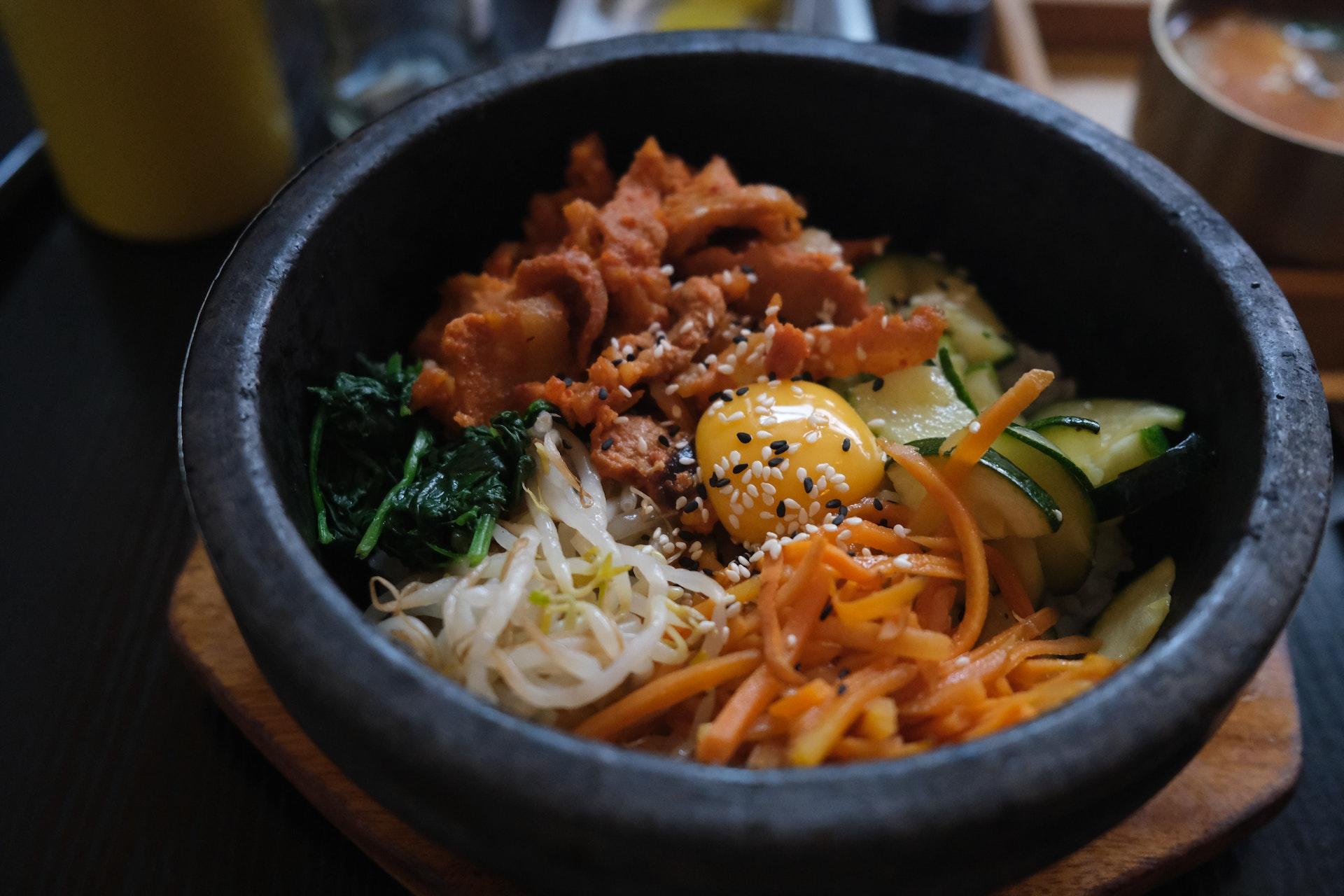 Preparado coreano em recipiente preto, visto de cima. Pode-se observar macarrão branco e de batata, carne, vegetais e outros ingredientes. Tudo está polvilhado com gergelim, algo muito comum na culinária local.