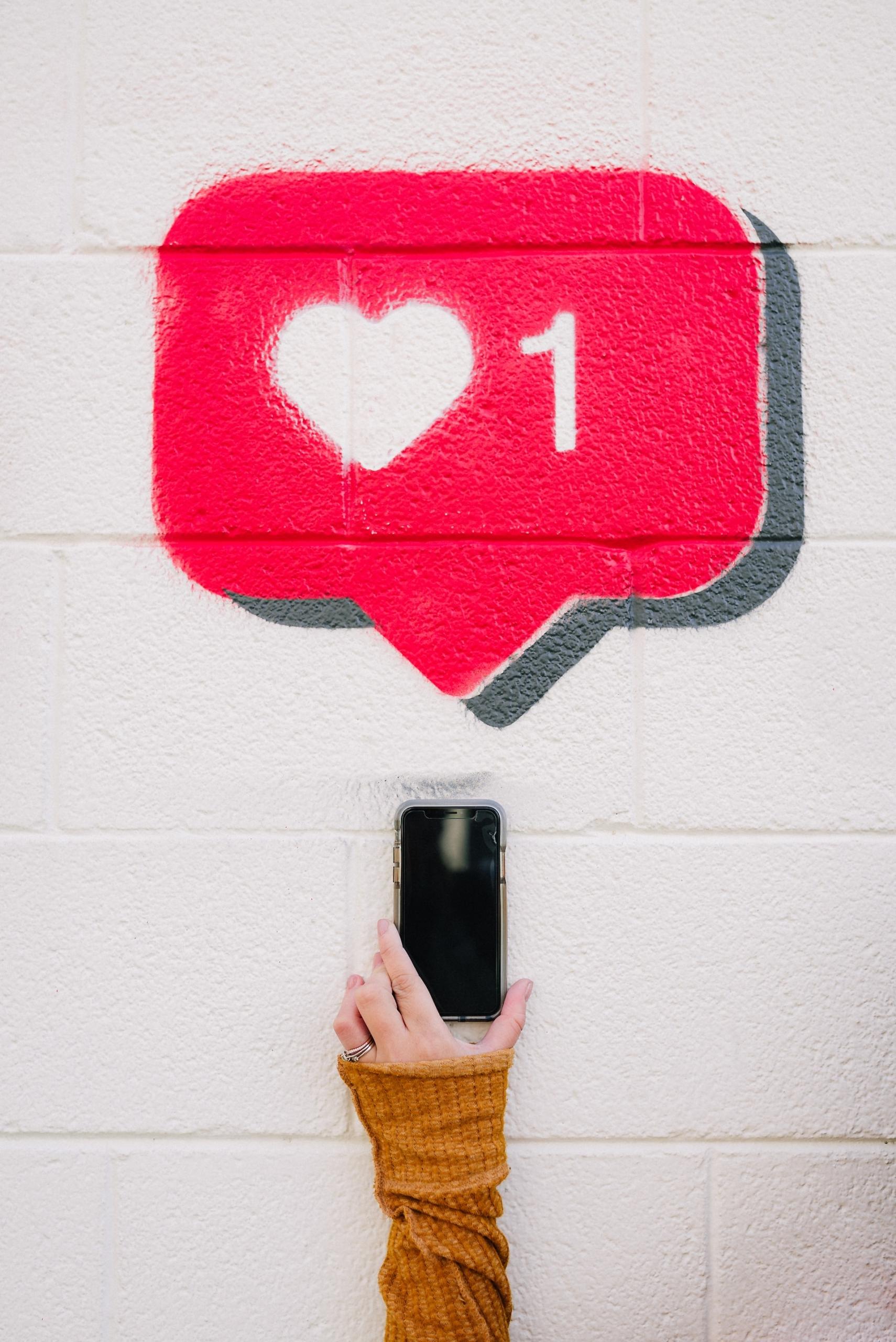 Mão com um celular em uma parede com um símbolo de like do Instagram pintado.