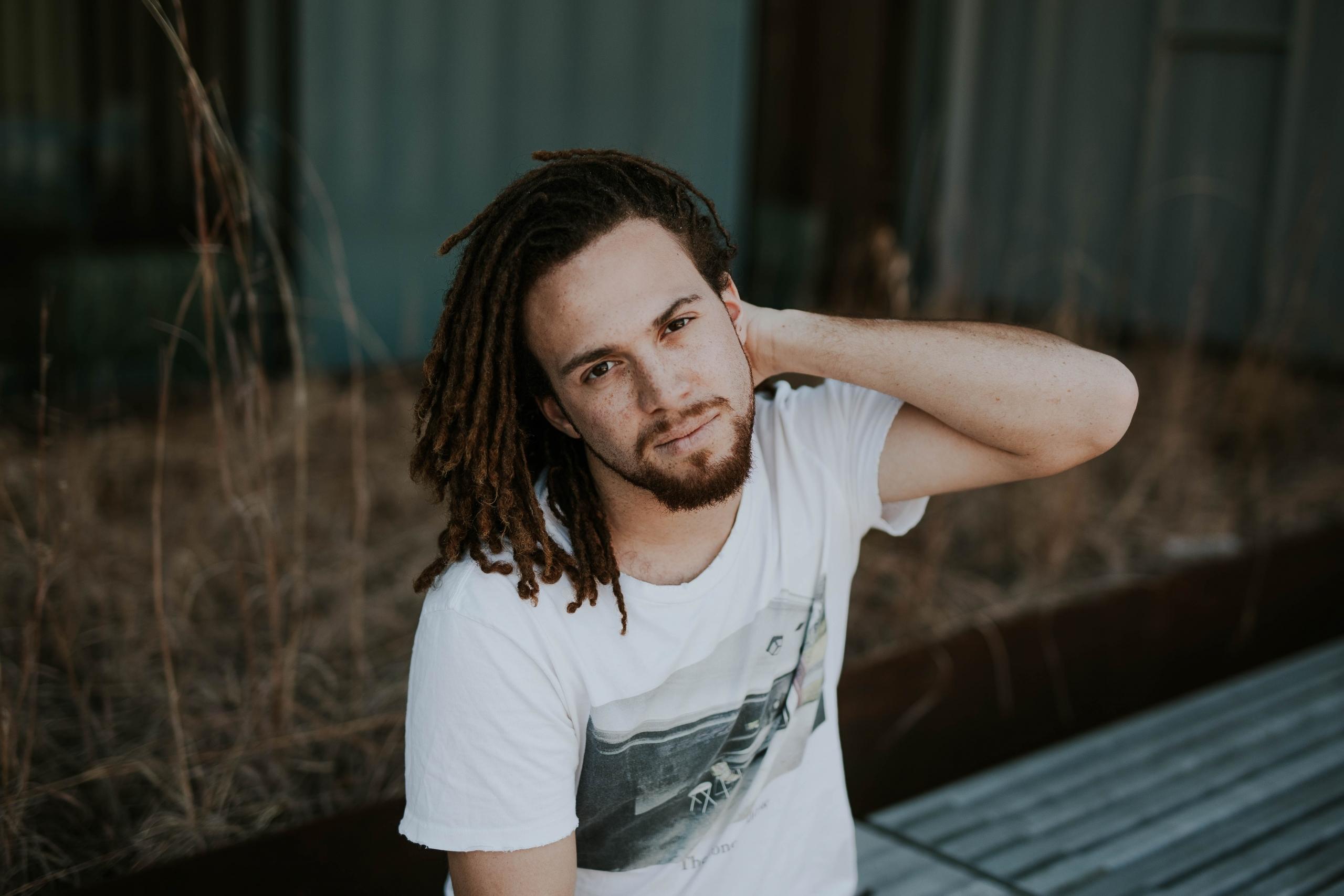 Homem branco utilizando tranças com camiseta clara e mão no pescoço