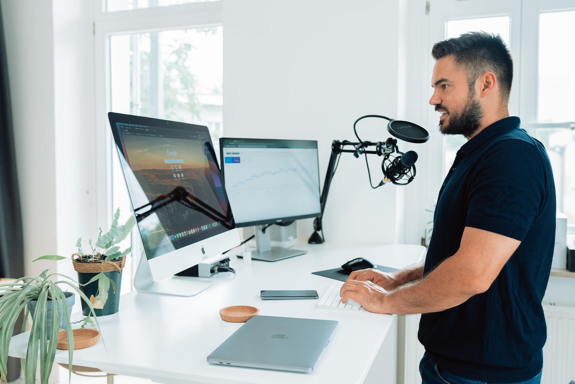 Homem em frente a dois monitores analisando um gráfico e com um microfone.