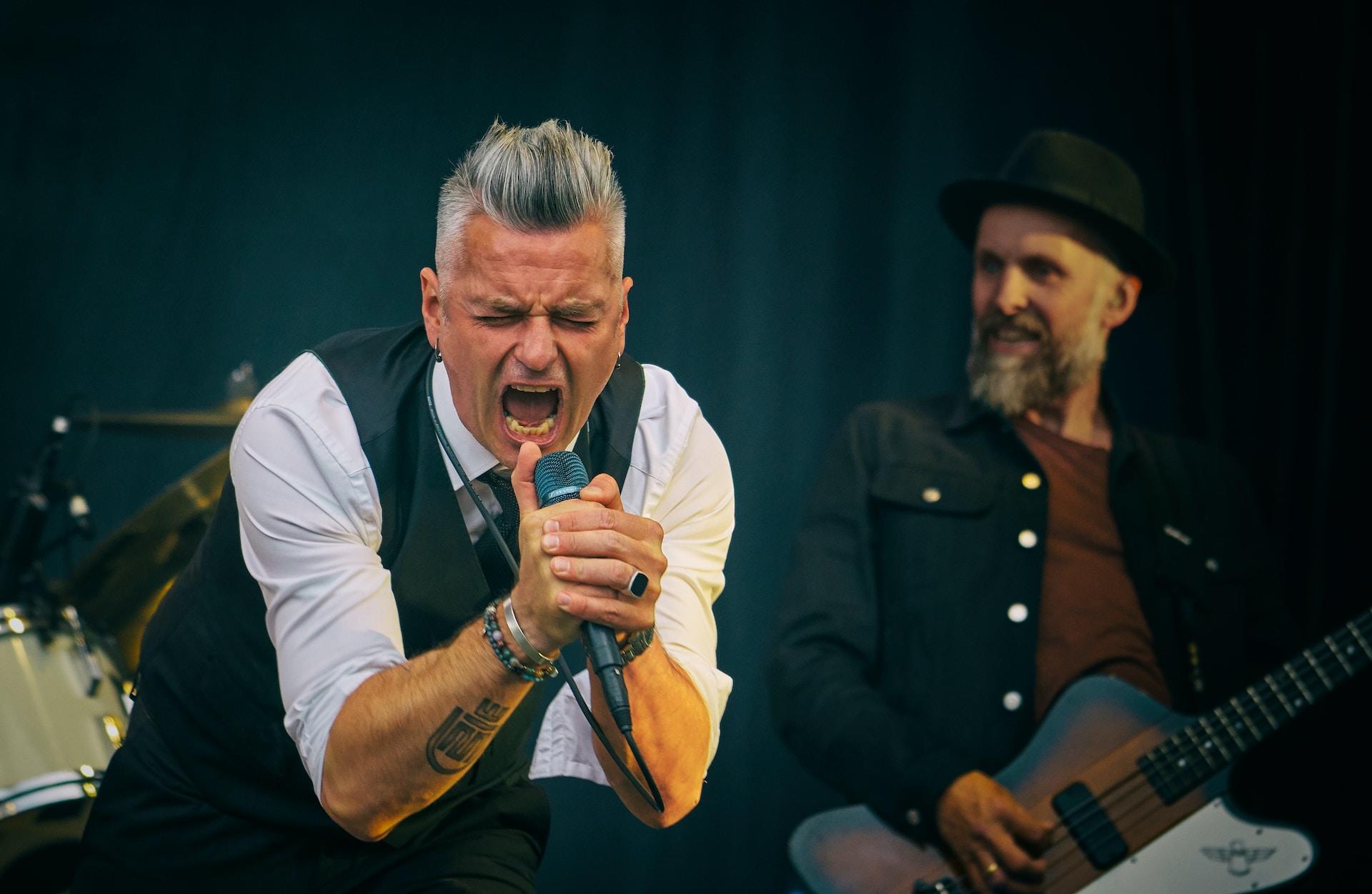 Dois homens apresentando em um palco, um deles cantando em um microfone e outro mais ao fundo tocando guitarra.