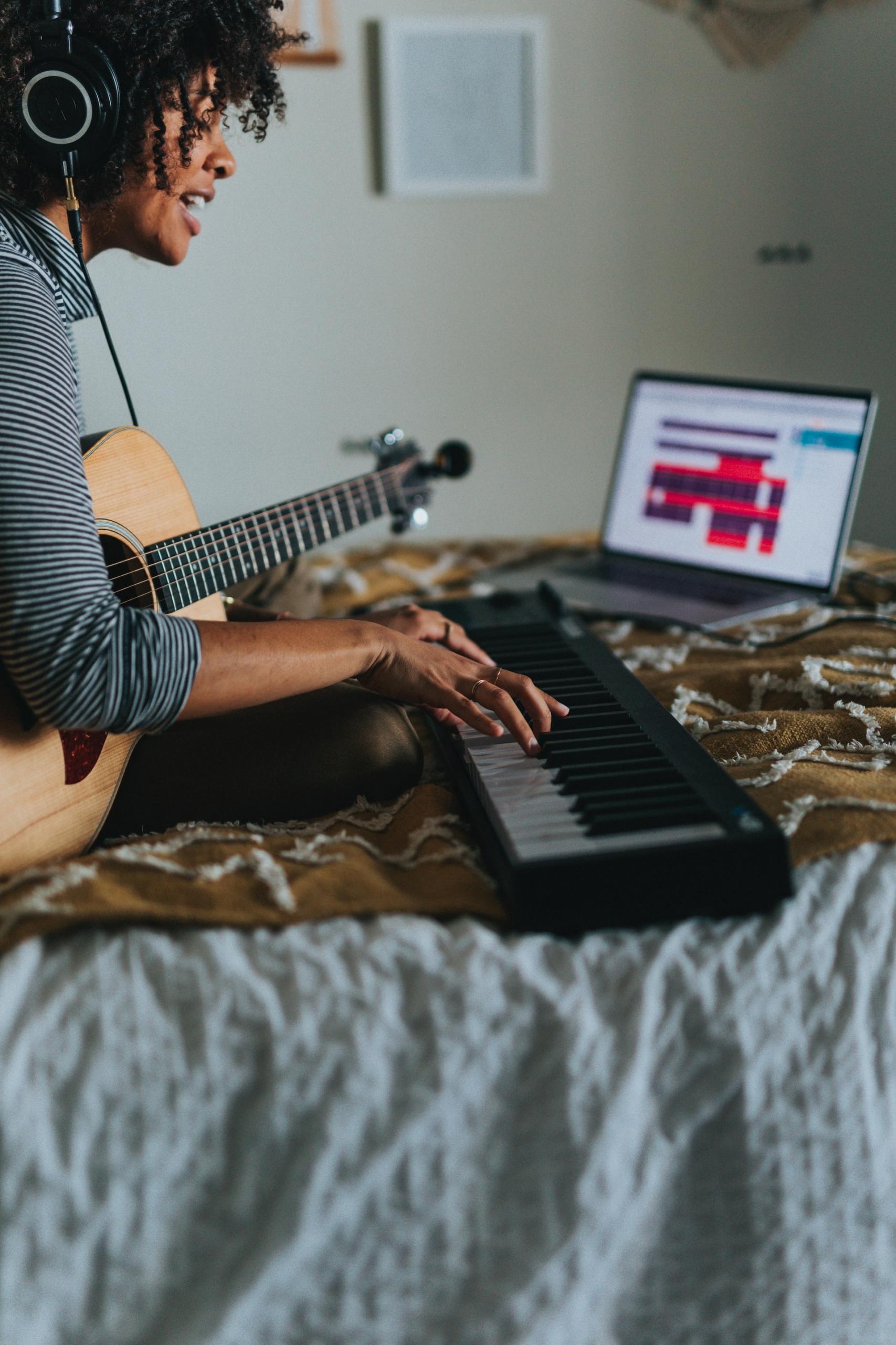 Mulher tocando teclaro e violão en cima de uma cama, com um notebook na frente.