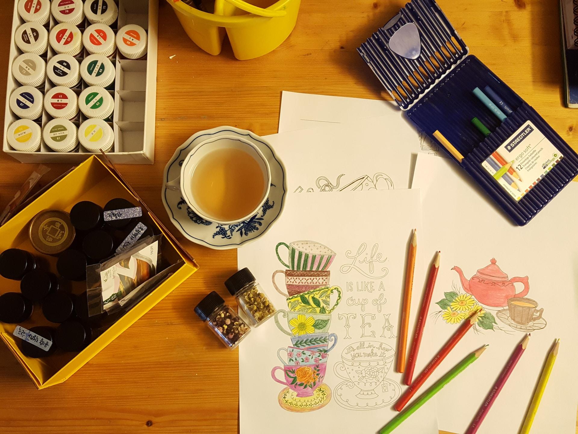 Foto de uma mesa com um desenho, uma xícara de chá e alguns lápis.