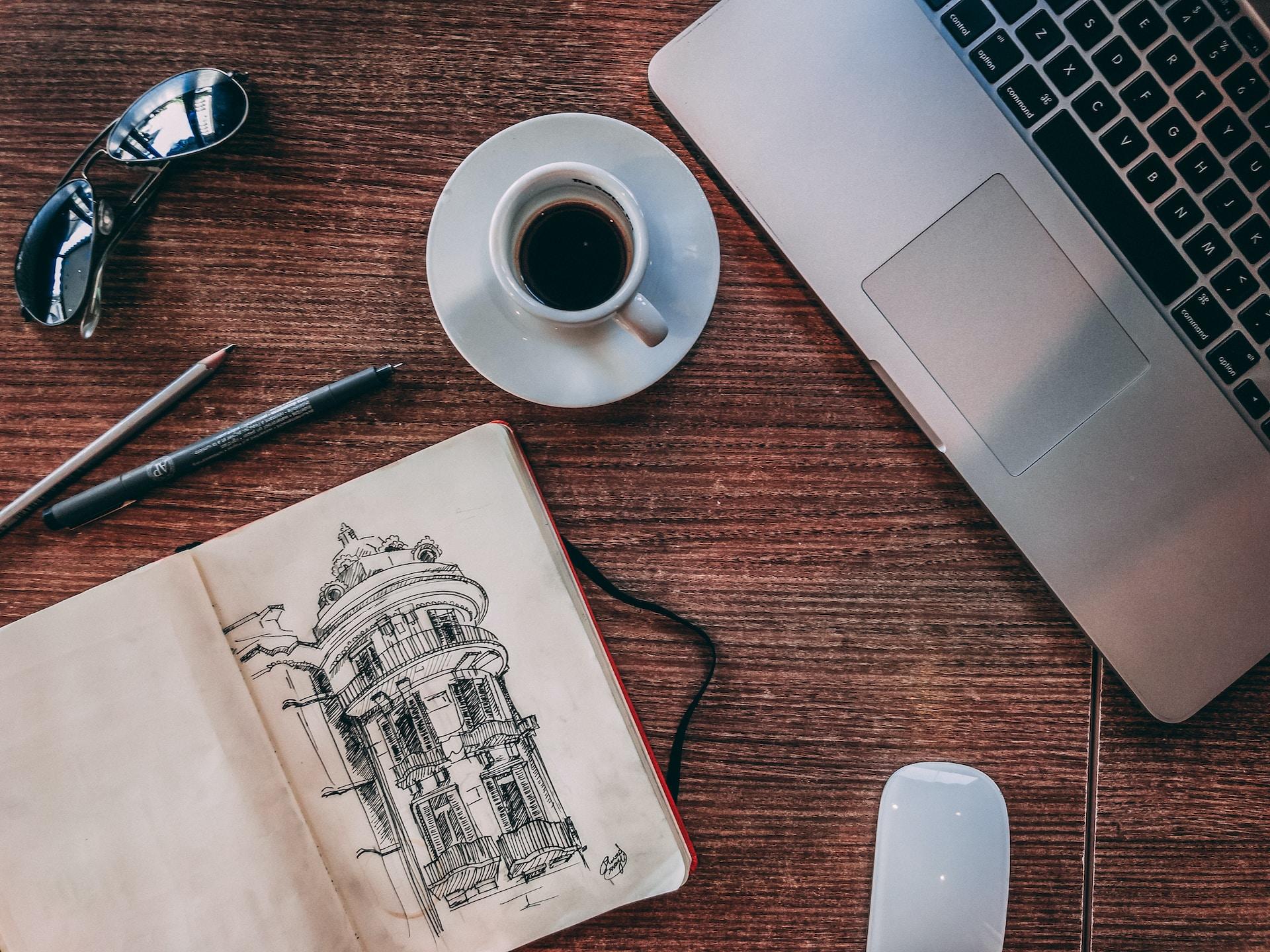 Fodo de uma mesa com um computador, um caderno com o desenho de um prédio, um xícara de café e um óculos de sol.