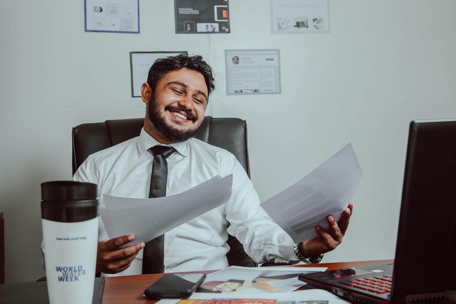 Homem em uma mesa de escritório, sorrindo e segurando alguns papéis.
