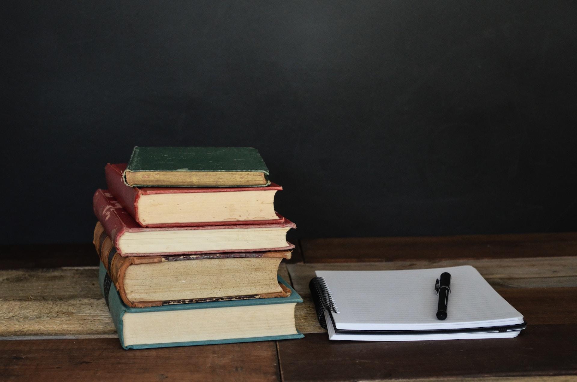 Livros empilhados em uma mesa com um caderno e caneta ao lado