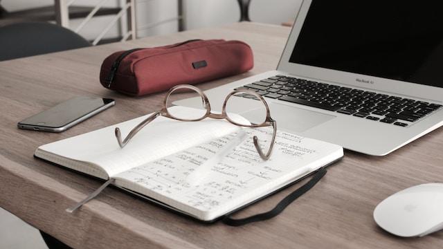 Caderno com óculos em cima e notebook do lado em uma mesa de madeira