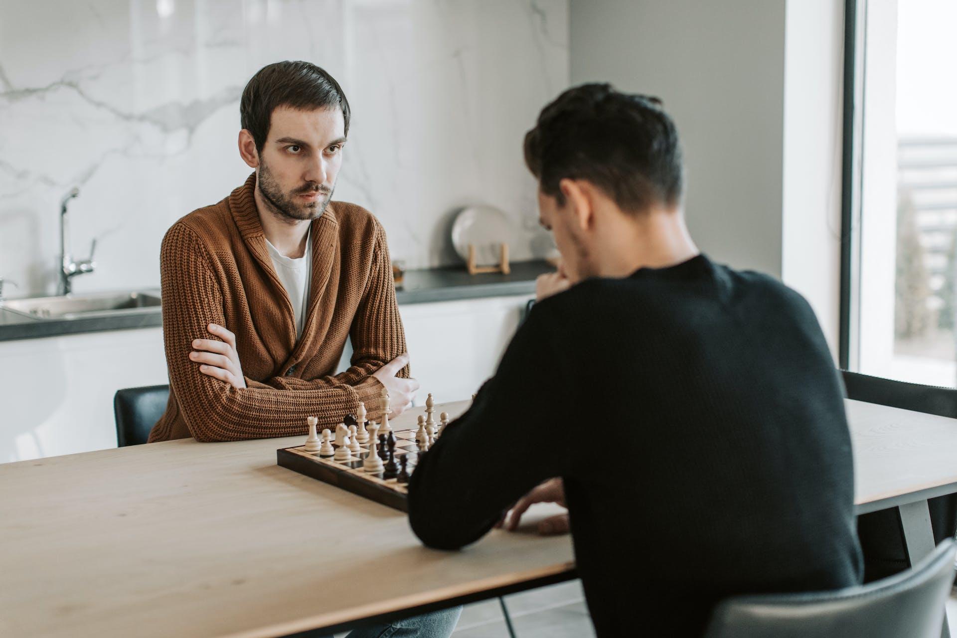 Dois rapazes sentados à mesa jogando xadrez. Um está de costas e veste preto. O outro está de frente e veste marrom. Este tem um semblante sério e concentrado.