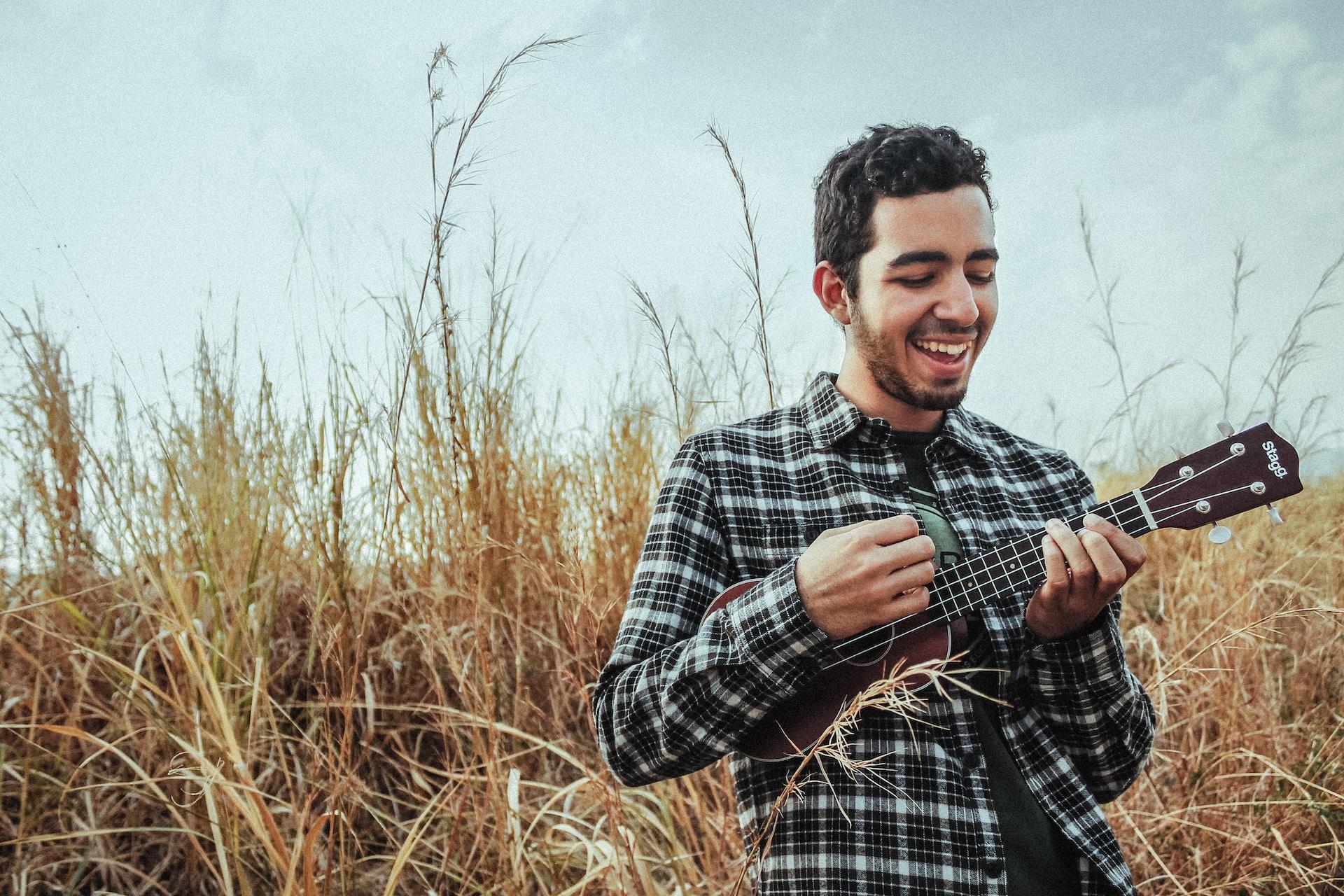 Homem sorrindo e tocando cavaquinho no meio de um mato seco.