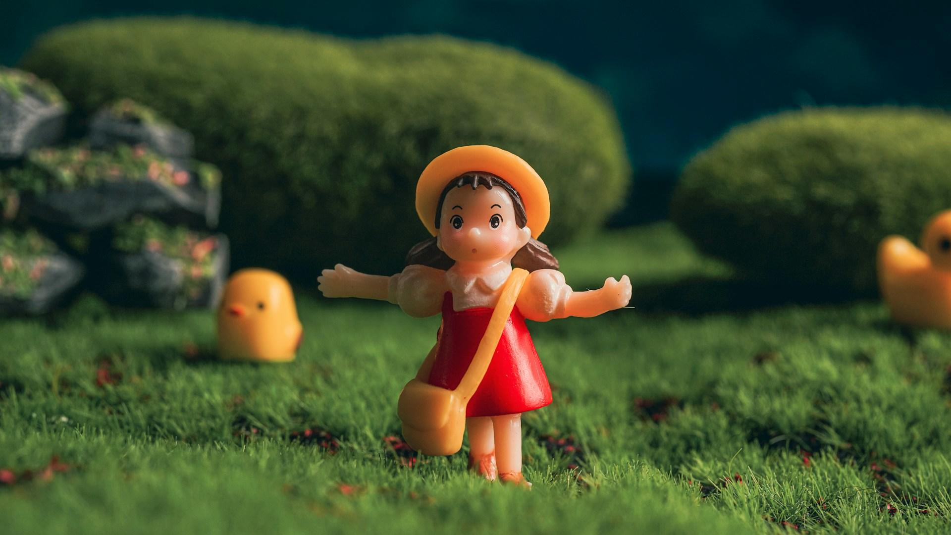 Foto de um boneco de uma menina em um cenário de grama com alguns patinhos ao fundo.