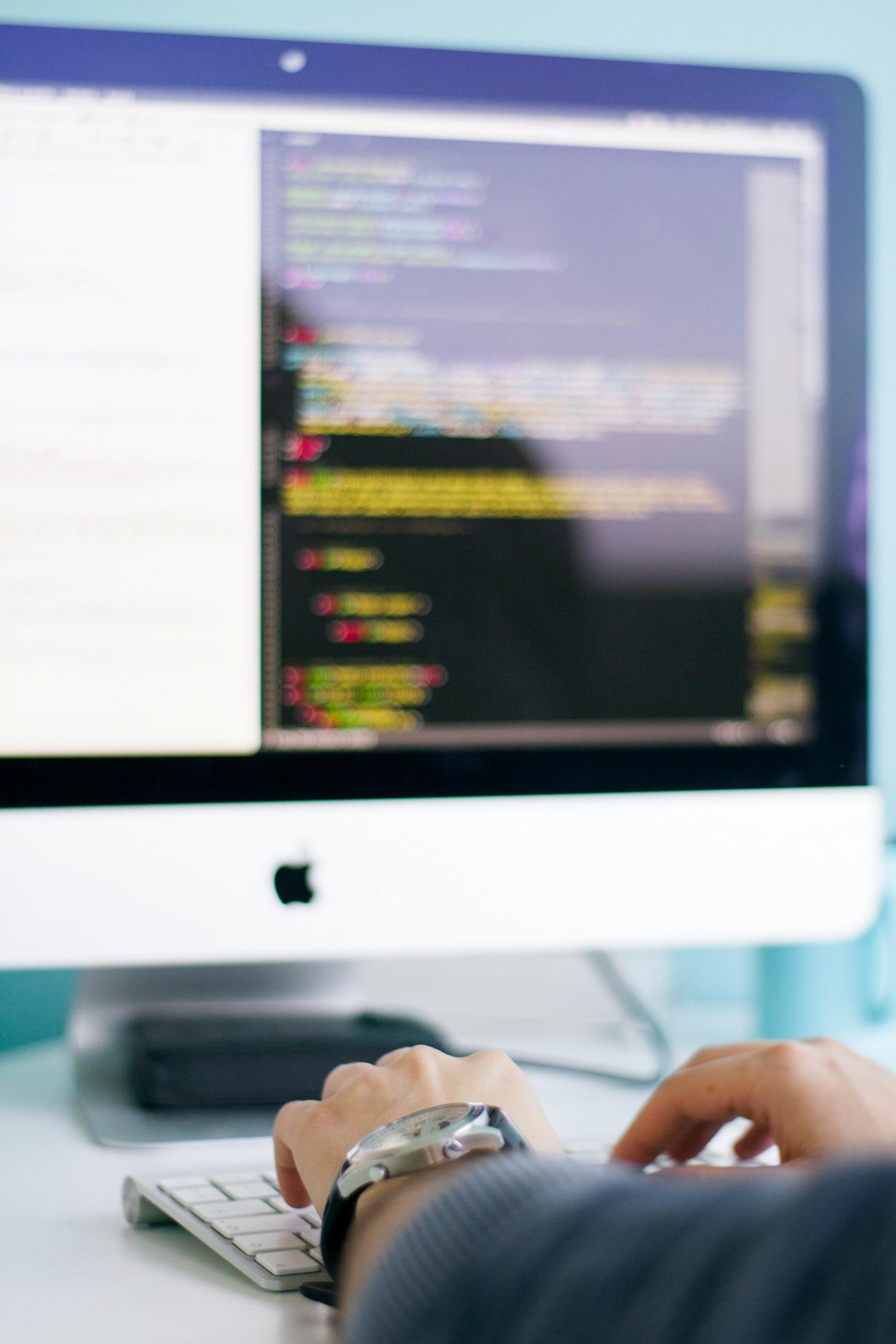 Imagem de mãos digitando em um teclado e a tela de um computador com códigos de programação.